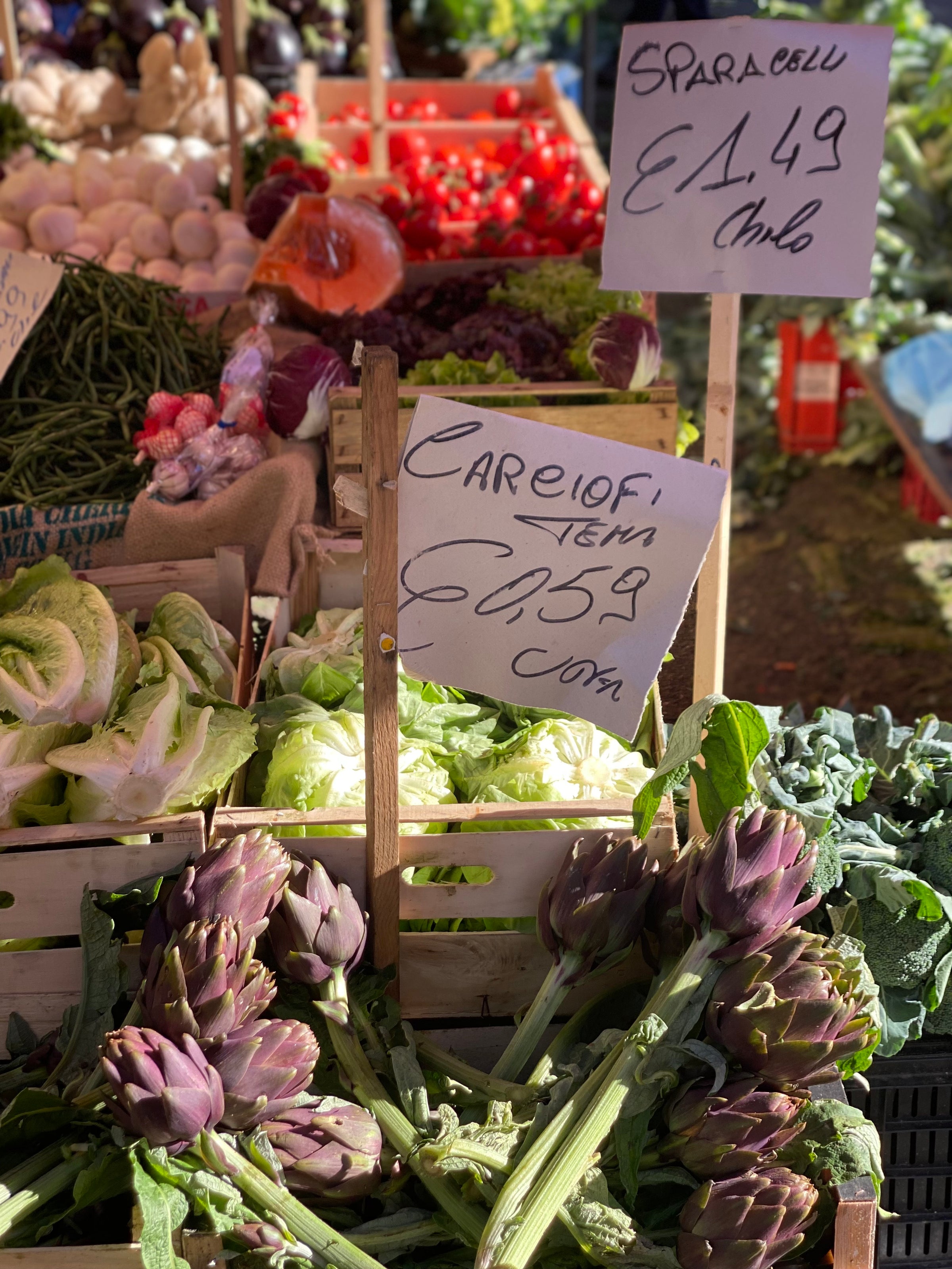 Fresh produce at an outdoor market, including artichokes, lettuce, tomatoes, and green beans. Handwritten price signs in Italian are visible among the vegetables.