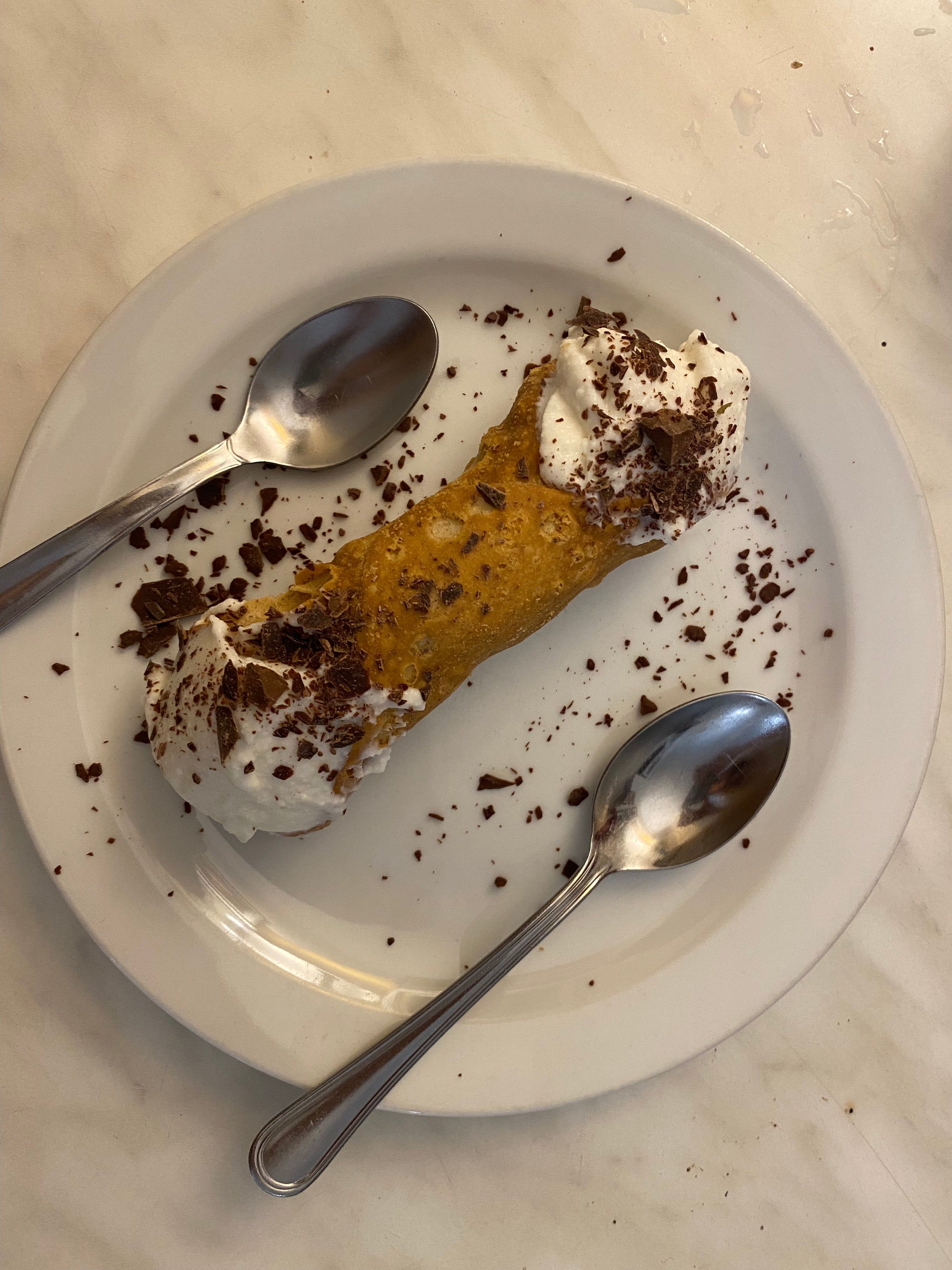 A single cannoli topped with whipped cream and chocolate shavings sits on a white plate, with two metal spoons placed on either side, on a marble surface.