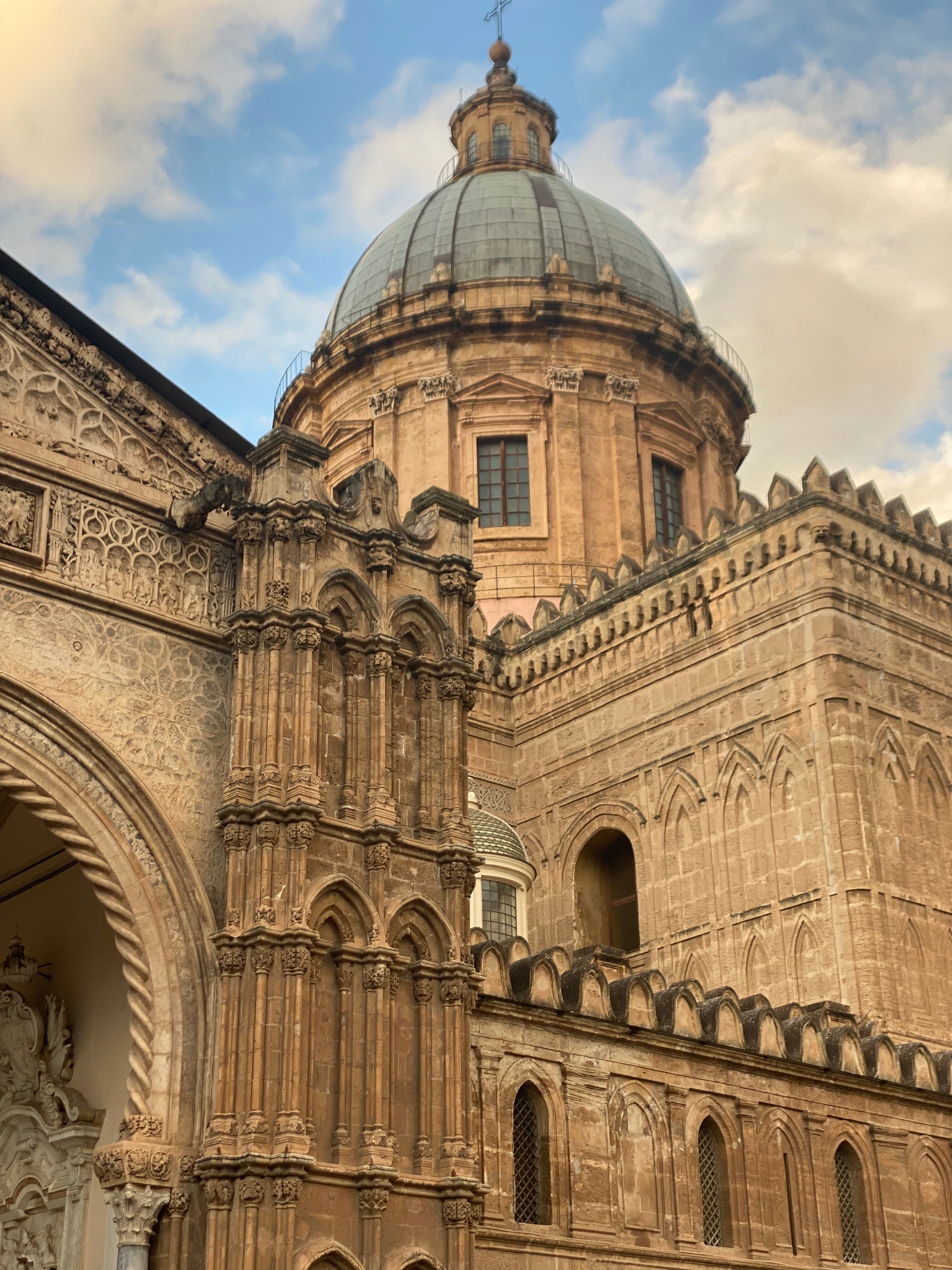 A close-up view of the ornate, tan stone facade and large dome of Palermo Cathedral in Sicily, Italy, set against a partly cloudy sky. The architecture shows elaborate carvings and Gothic details.