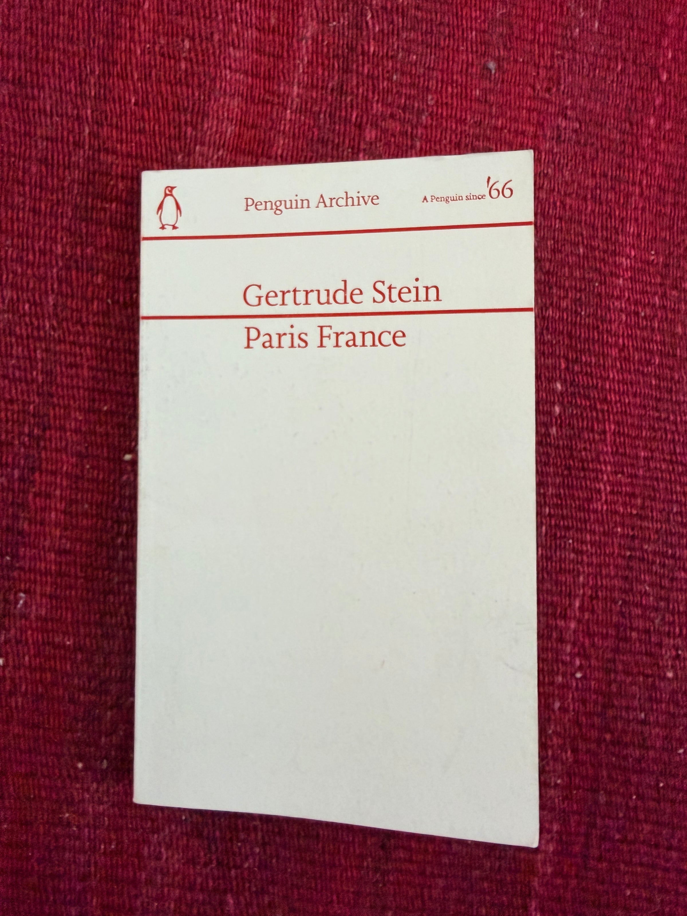 A white book titled Gertrude Stein Paris France from the Penguin Archive series lies on a textured red surface.