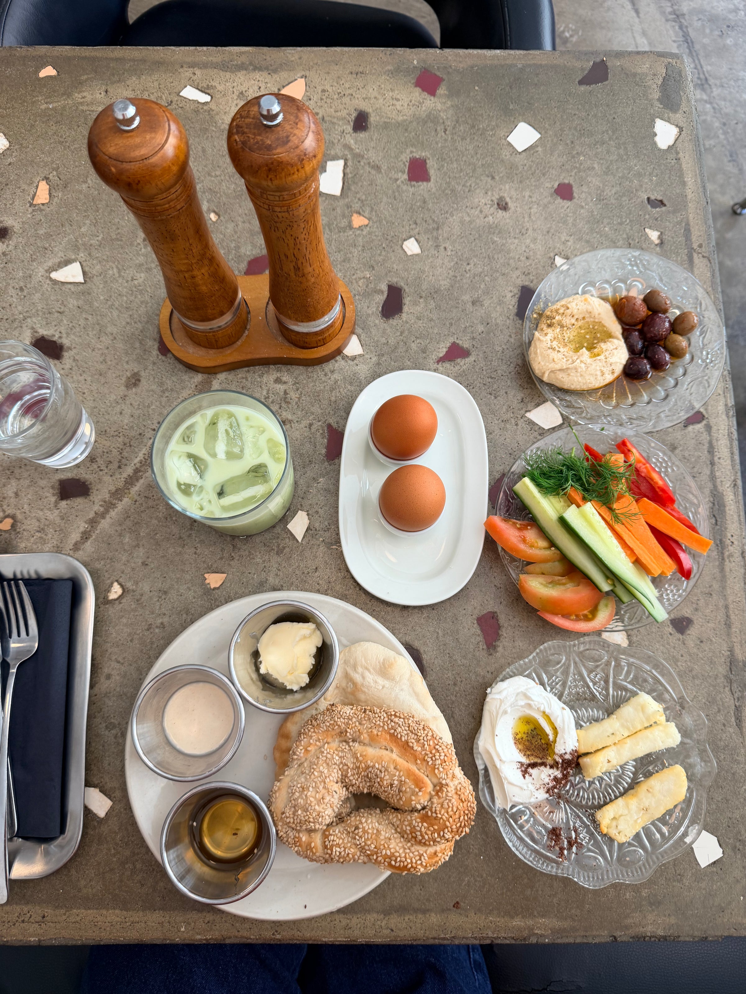 A Mediterranean breakfast on a stone table includes eggs, bread with sesame seeds, dips, butter, fresh vegetables, cheese, olives, hummus, a green drink with ice, salt and pepper shakers, and cutlery in a napkin.