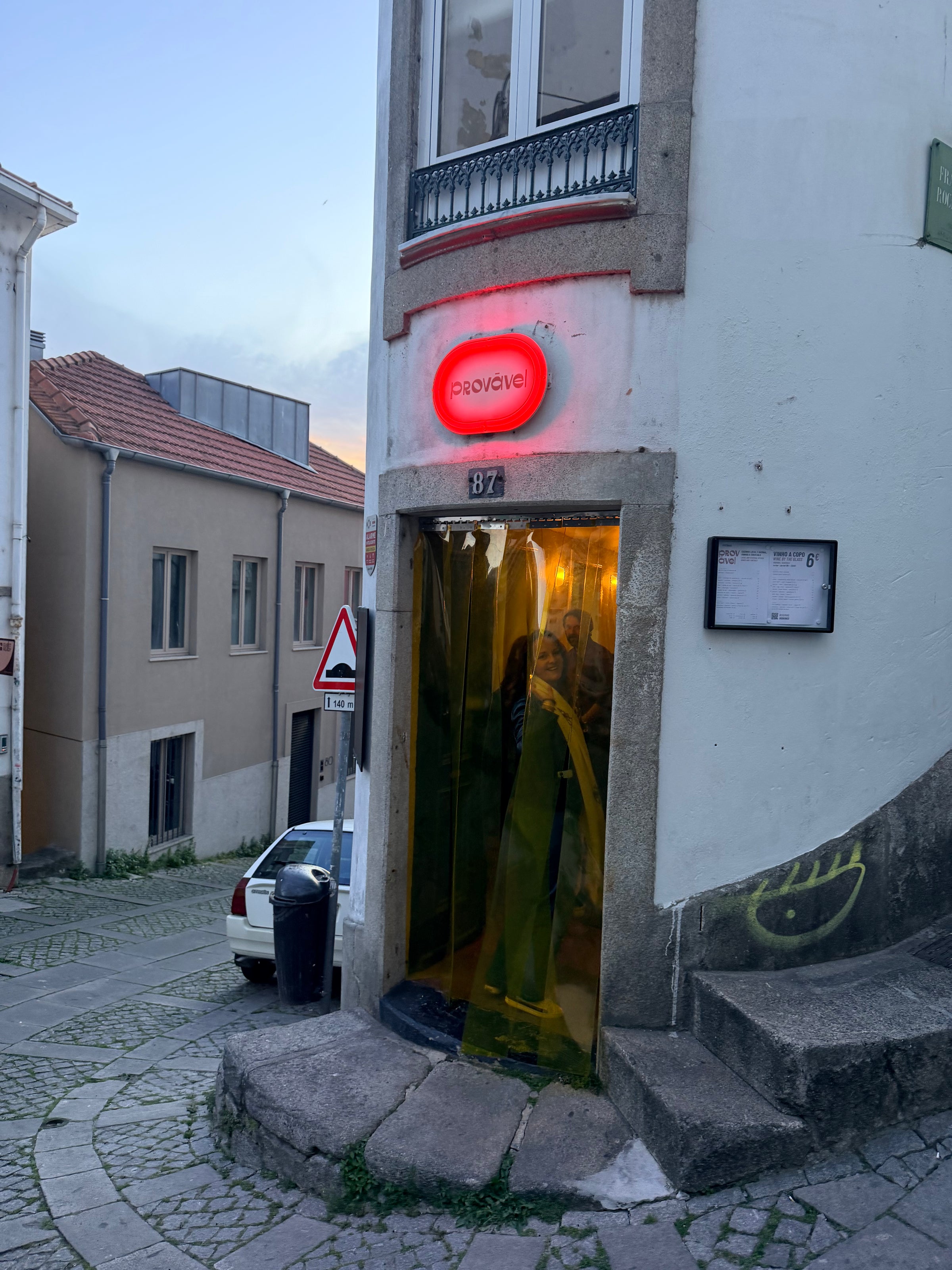 A narrow corner building with a glowing red PROVAÇO sign above a yellow-lit entrance at dusk, located on a sloped cobblestone street with a trash bin and street signs nearby.