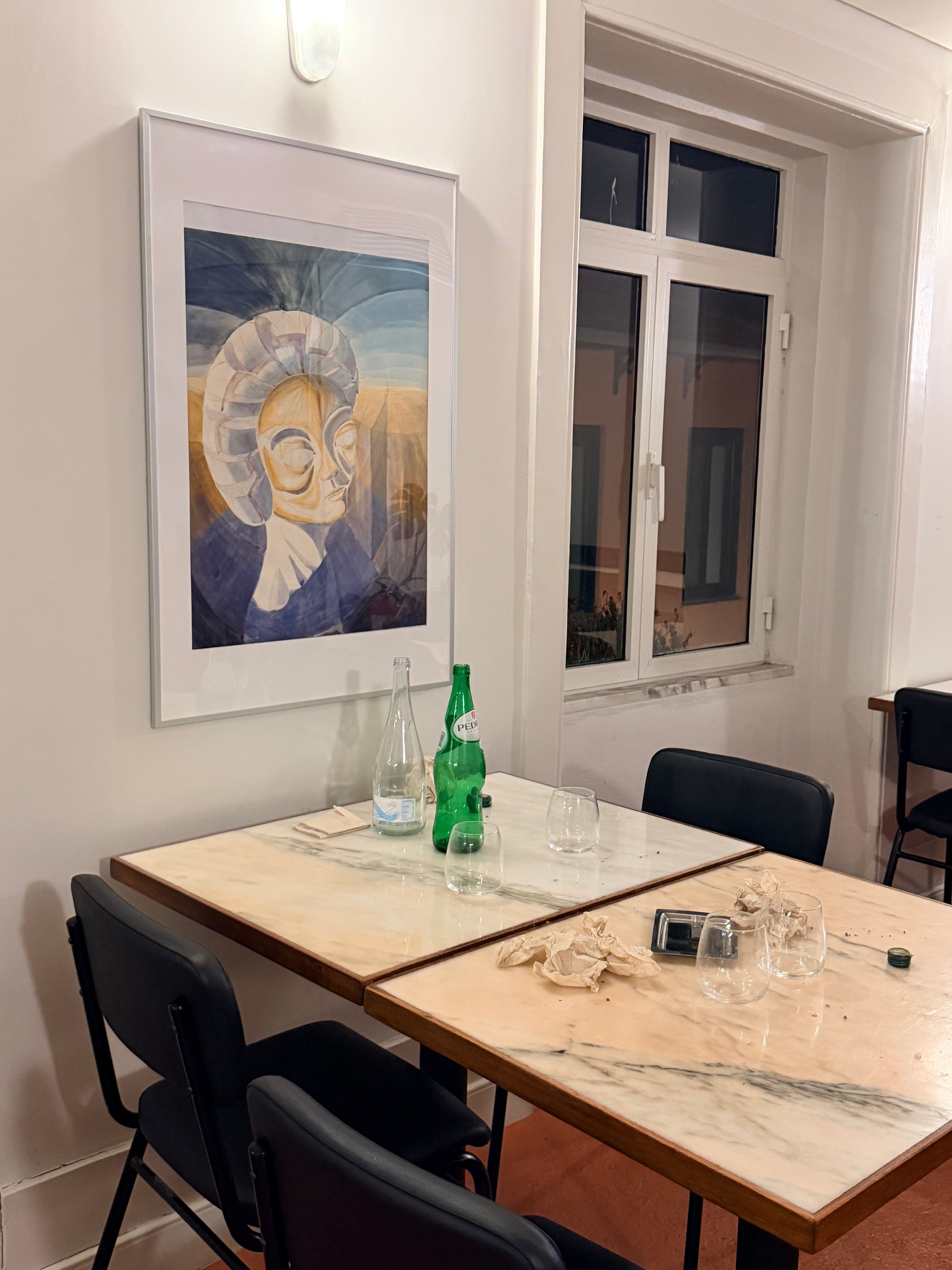 Two marble tables with black chairs are set with empty glasses, bottles, and a phone. A framed abstract painting of a figure hangs on a white wall, and a window reveals a nighttime scene outside.