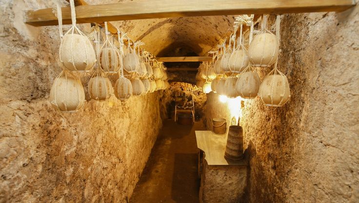 A stone-walled cellar with arched ceilings, filled with round cheese wheels hanging from ropes. A wooden table and barrels are visible under warm lighting at the end of the narrow, rustic corridor.