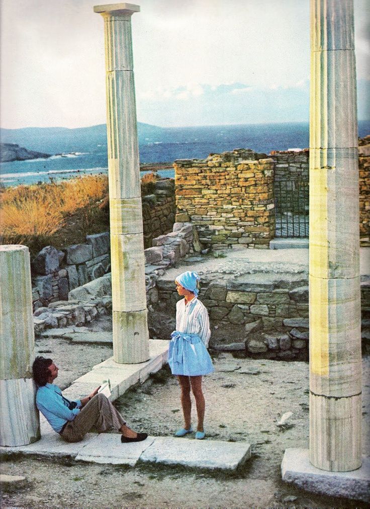 A man sits and sketches while a woman in a light blue skirt and hat stands nearby among ancient stone ruins with tall columns, against a backdrop of sea and hills.