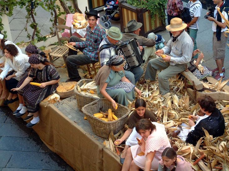 A group of people in traditional clothing sit and work with corn, shelling cobs and gathering husks, while a man plays an accordion. Large baskets of corn are on the table, and some onlookers stand nearby.