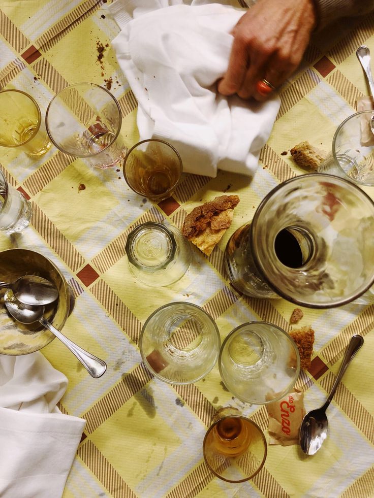 A cluttered table with empty glasses, spoons, crumbs, and a glass carafe on a yellow checkered tablecloth. A hand holding a napkin is wiping the table.