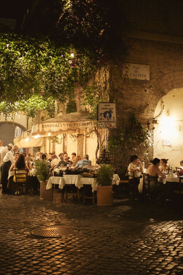 A cozy outdoor Italian restaurant at night with people dining under warm lights and greenery, set on a cobblestone street beneath a sign reading Via di Calisto.