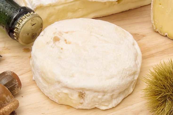 A round, creamy white cheese sits on a wooden board next to a bottle, a chestnut, acorns, and wedges of other cheeses. The cheese has a soft, wrinkled rind and a slightly uneven surface.