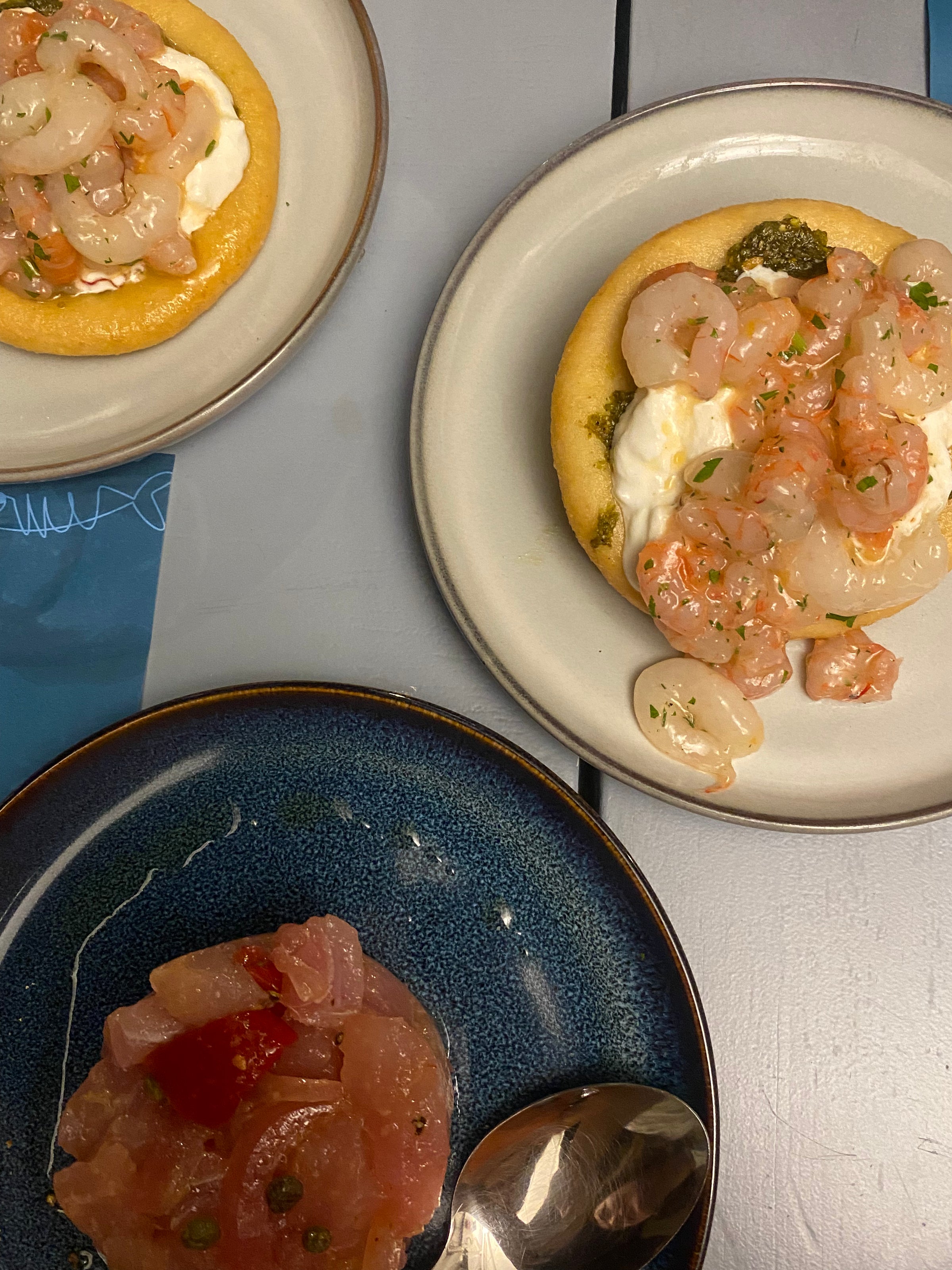 Three plates with seafood dishes: two plates have mini flatbreads topped with shrimp and cream, while the third plate has a serving of raw fish with capers and a spoon beside it. The plates sit on a gray and blue table.
