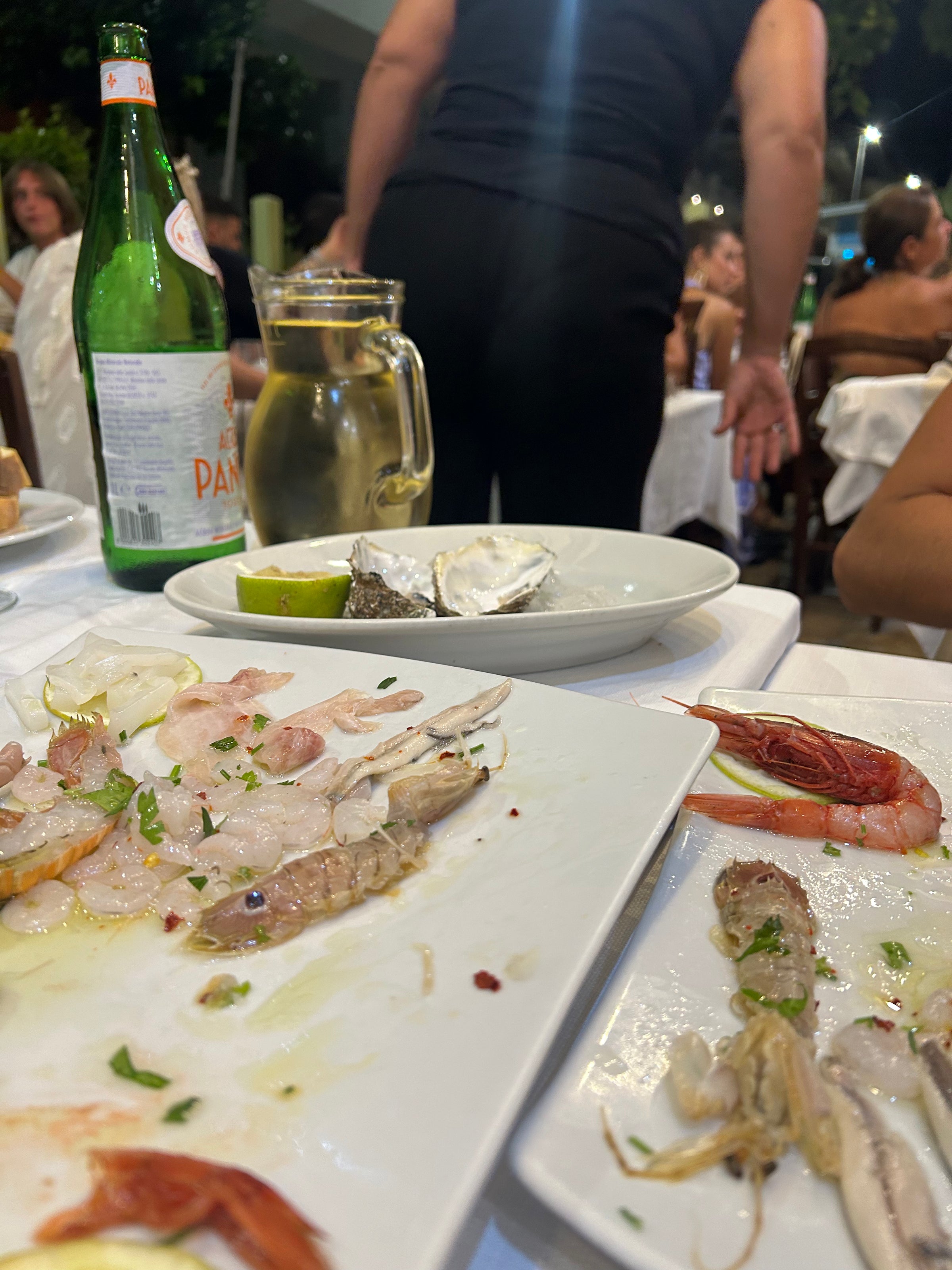Plates with assorted raw seafood, a red prawn, and empty oyster shells are on a table. A beer bottle, a jug of water, and diners sitting at surrounding tables are visible in the background.