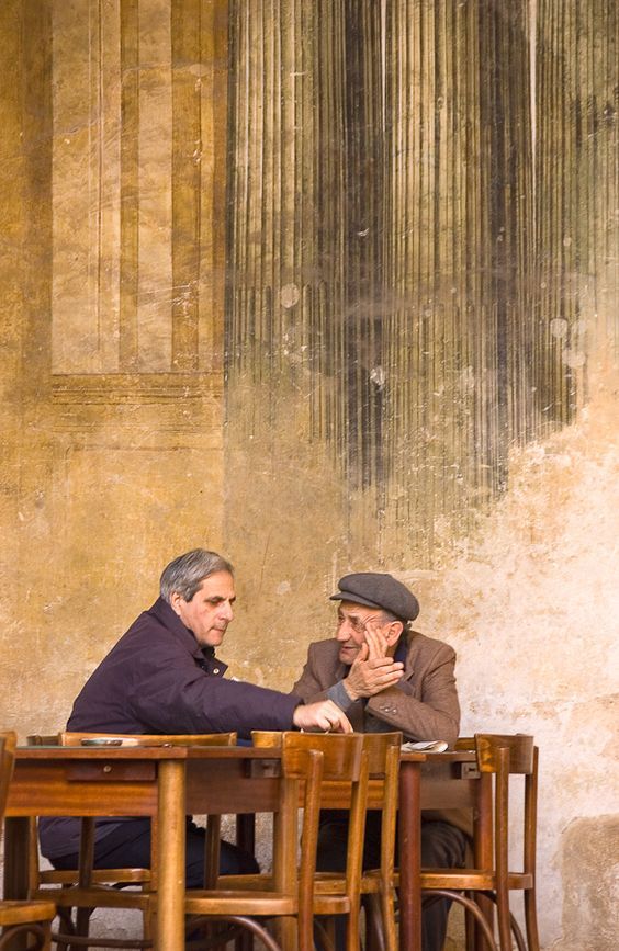 Two older men sit at a wooden table, engaged in conversation. One man smiles while wearing a hat, the other gestures with his hand. The background is a textured, aged wall with vertical streaks and faded architectural details.