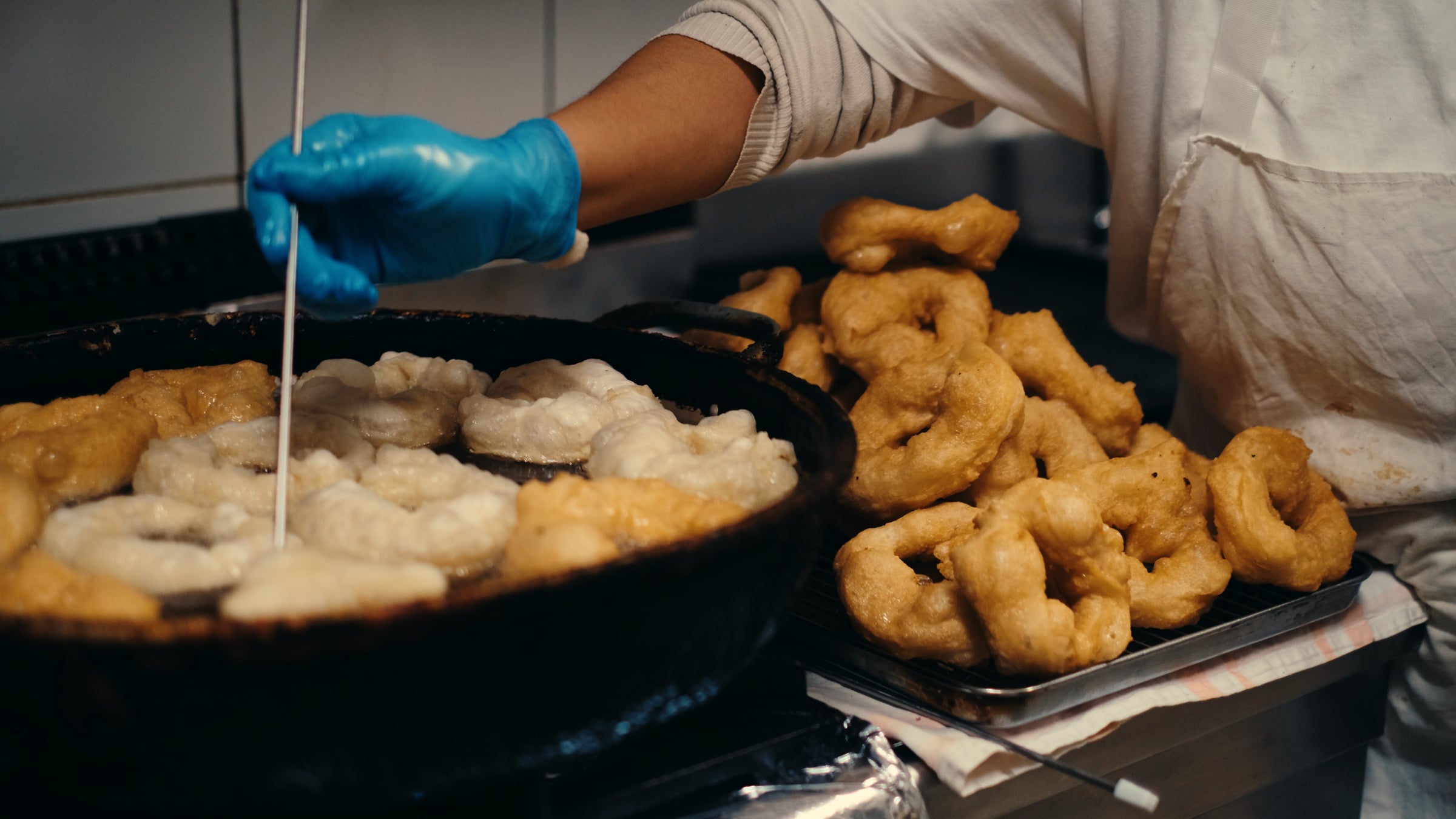 A person wearing a blue glove fries dough rings in a pan, while a pile of cooked golden doughnuts rests on a tray beside them.