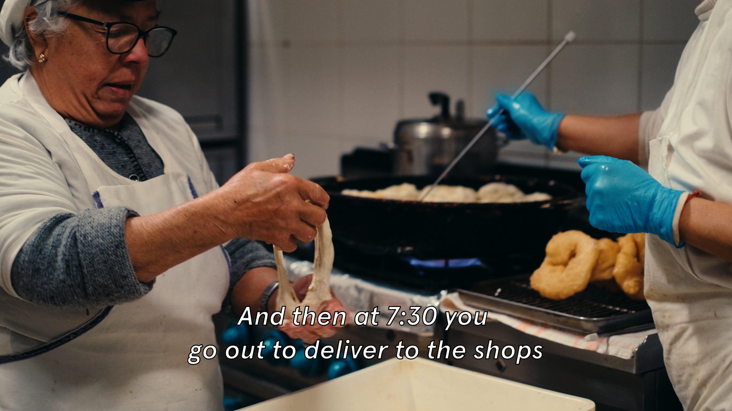 Two people in a kitchen prepare doughnuts. One shapes dough while the other, wearing blue gloves, uses a skimmer over hot oil. Text on image reads: And then at 7:30 you go out to deliver to the shops.