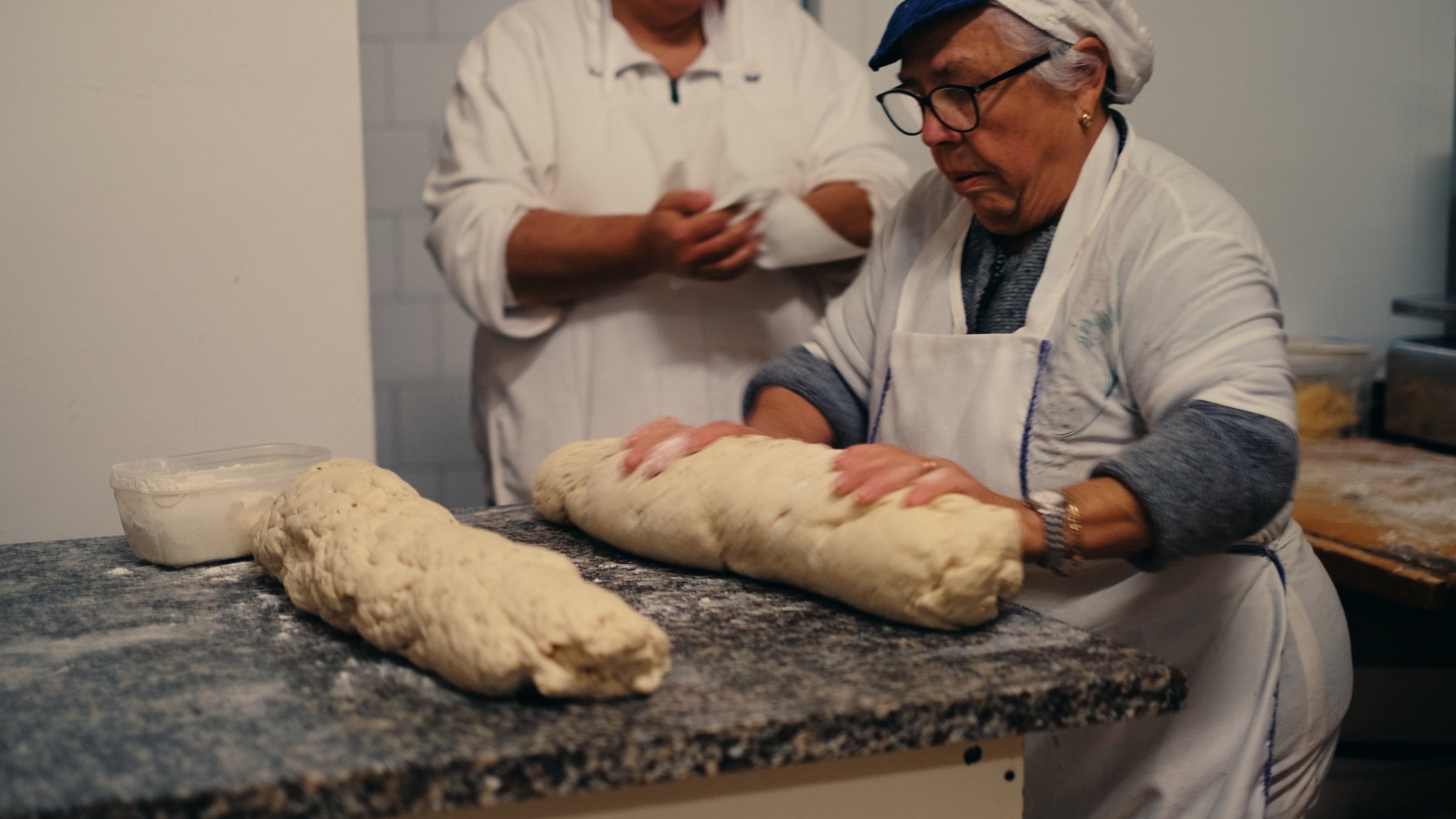 An elderly woman wearing glasses and a headscarf kneads a large piece of dough on a floured countertop, while another person in a white apron stands behind her, preparing to bake.