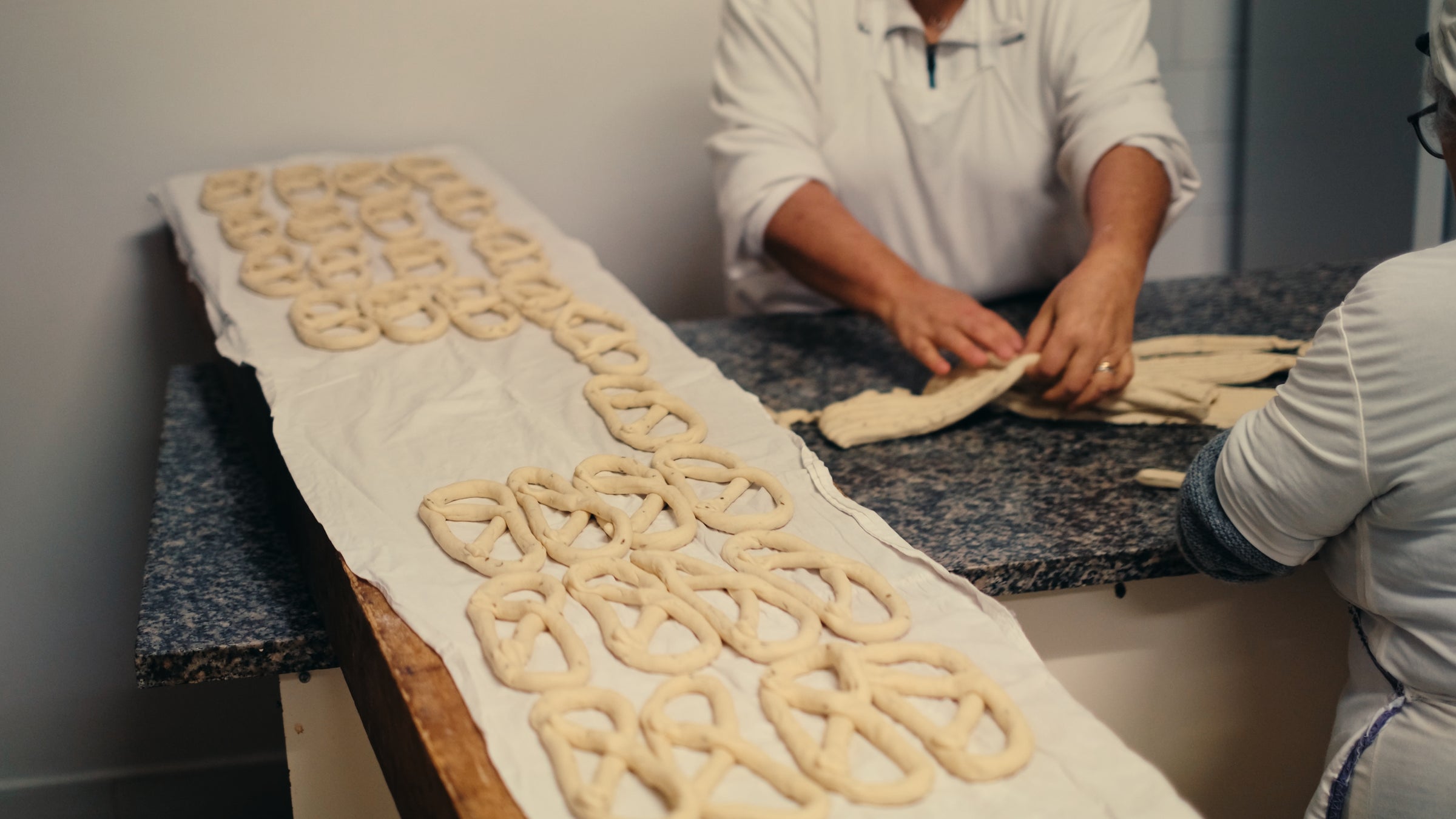 Two people shape pretzel dough by hand on a dark countertop. Rows of unbaked pretzels rest on a white cloth on a wooden table, ready for baking. The image focuses on the hands and dough, not showing faces.