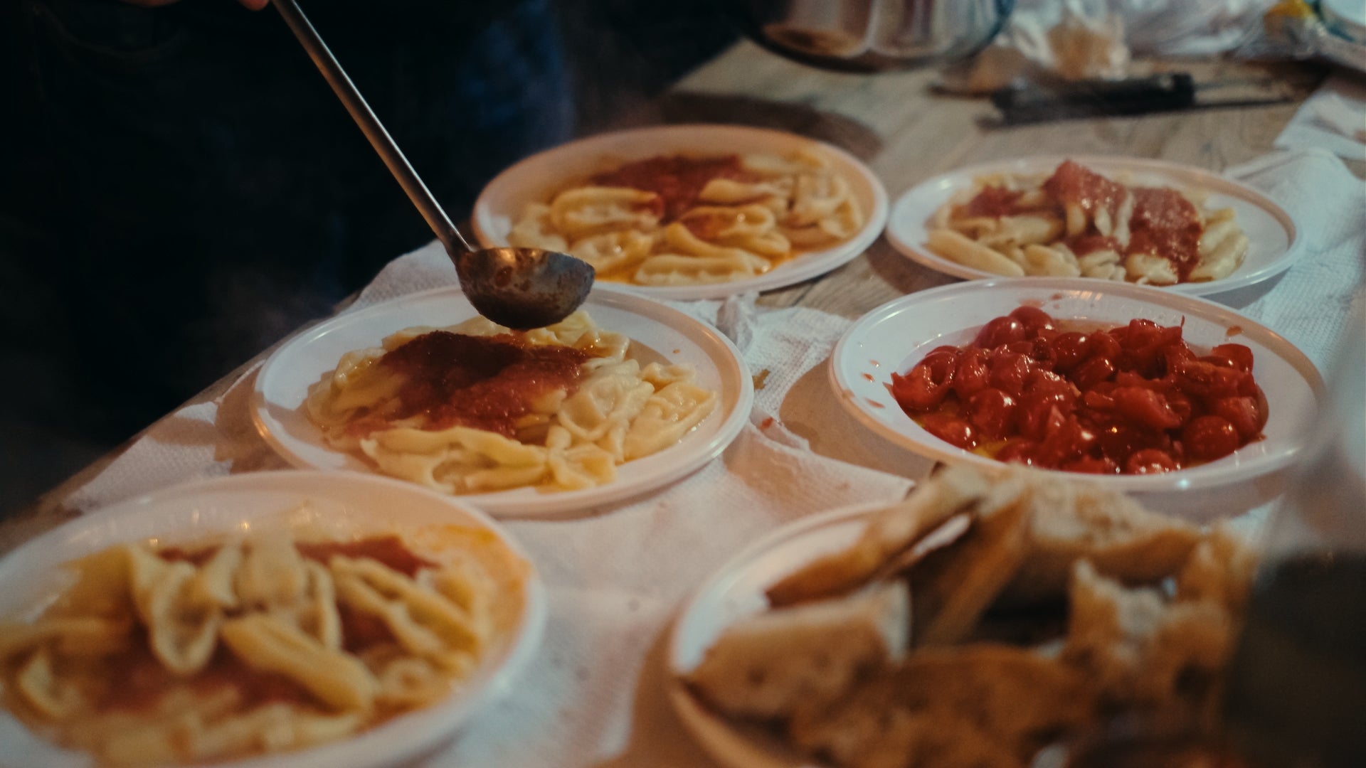 A person ladles tomato sauce onto plates of pasta, with several dishes of pasta and a bowl of cherry tomatoes arranged on a table covered with a white cloth. Pieces of bread are visible in the foreground.