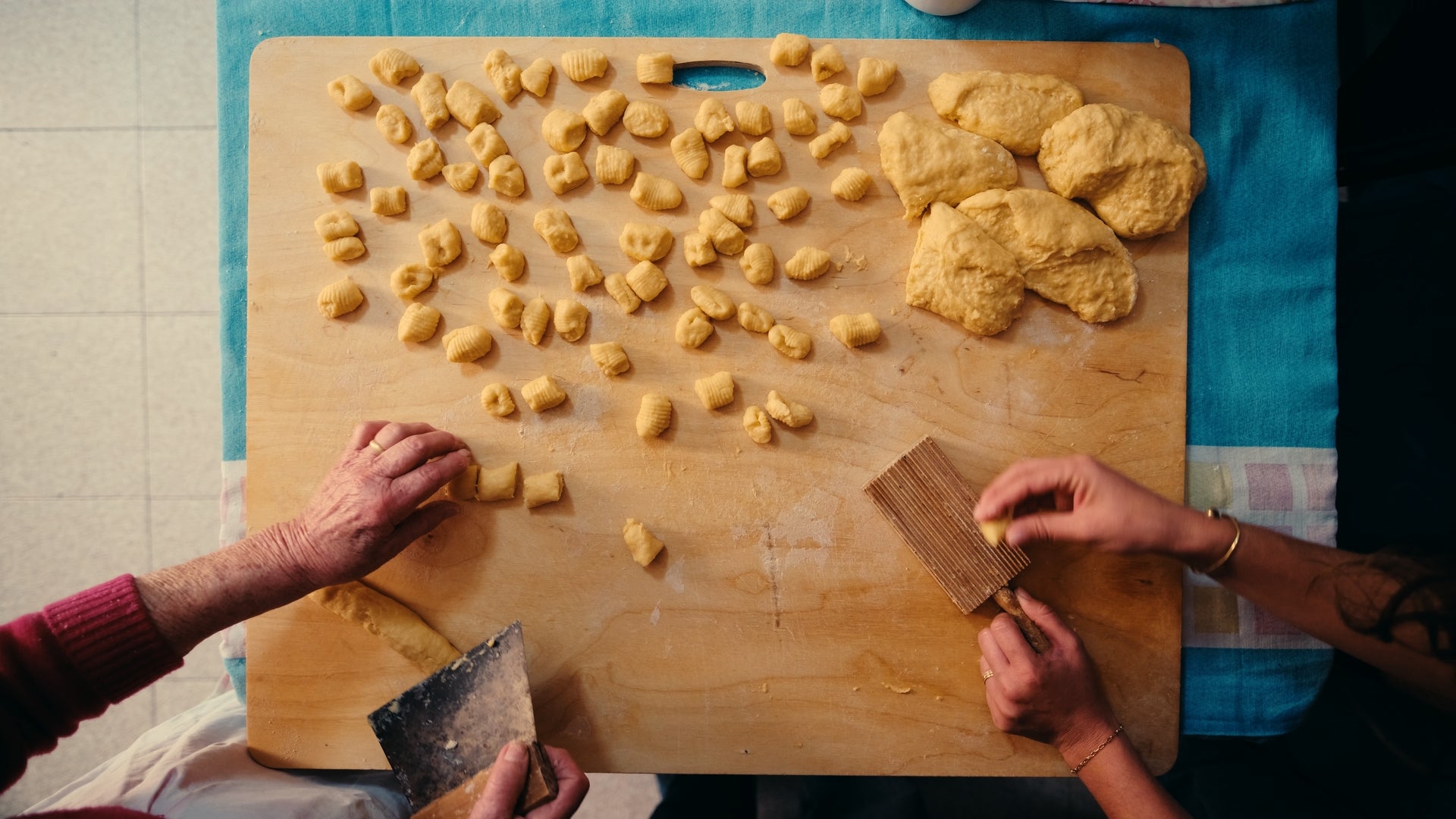 Two people shaping gnocchi dough on a wooden board. Several small dough pieces and rolled gnocchi are visible, with dough balls on the side. Hands are working with a ridged tool and a dough cutter.