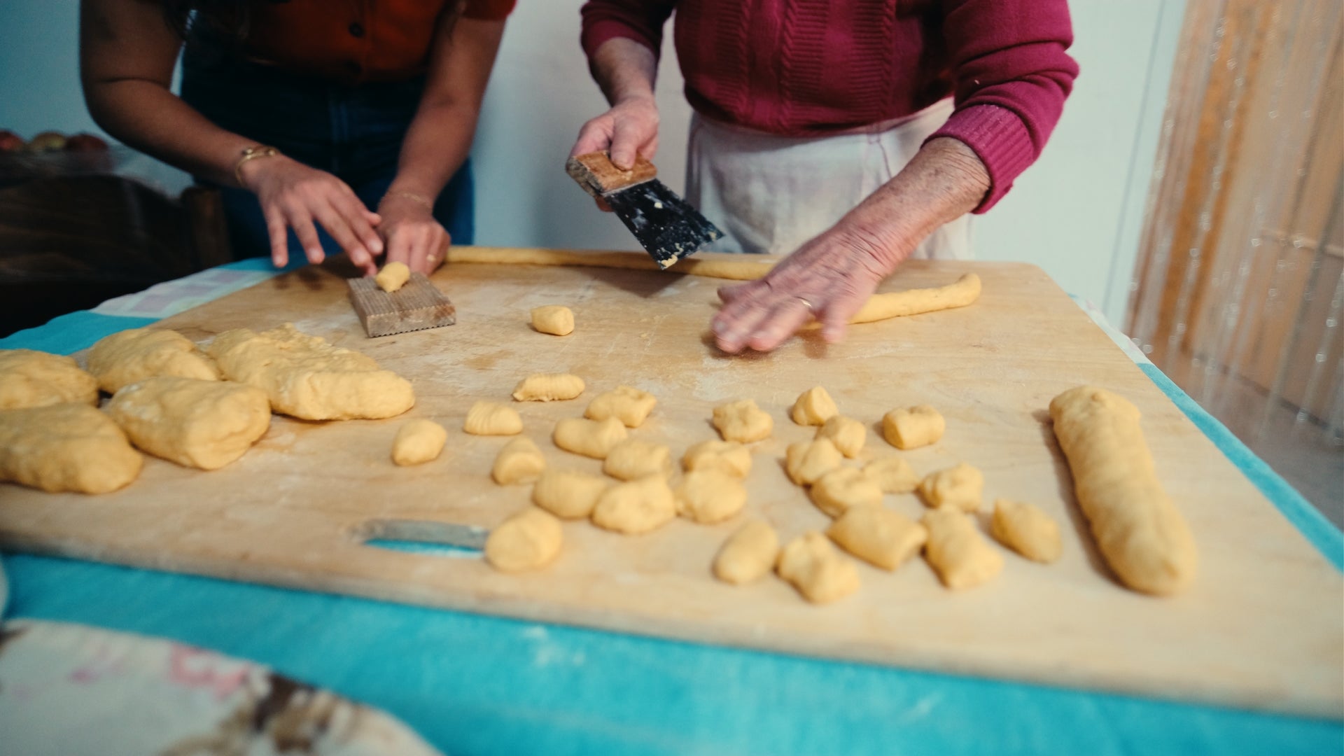 Two people work together shaping dough on a wooden board; there are small dough pieces scattered around and long rolls of dough being cut.