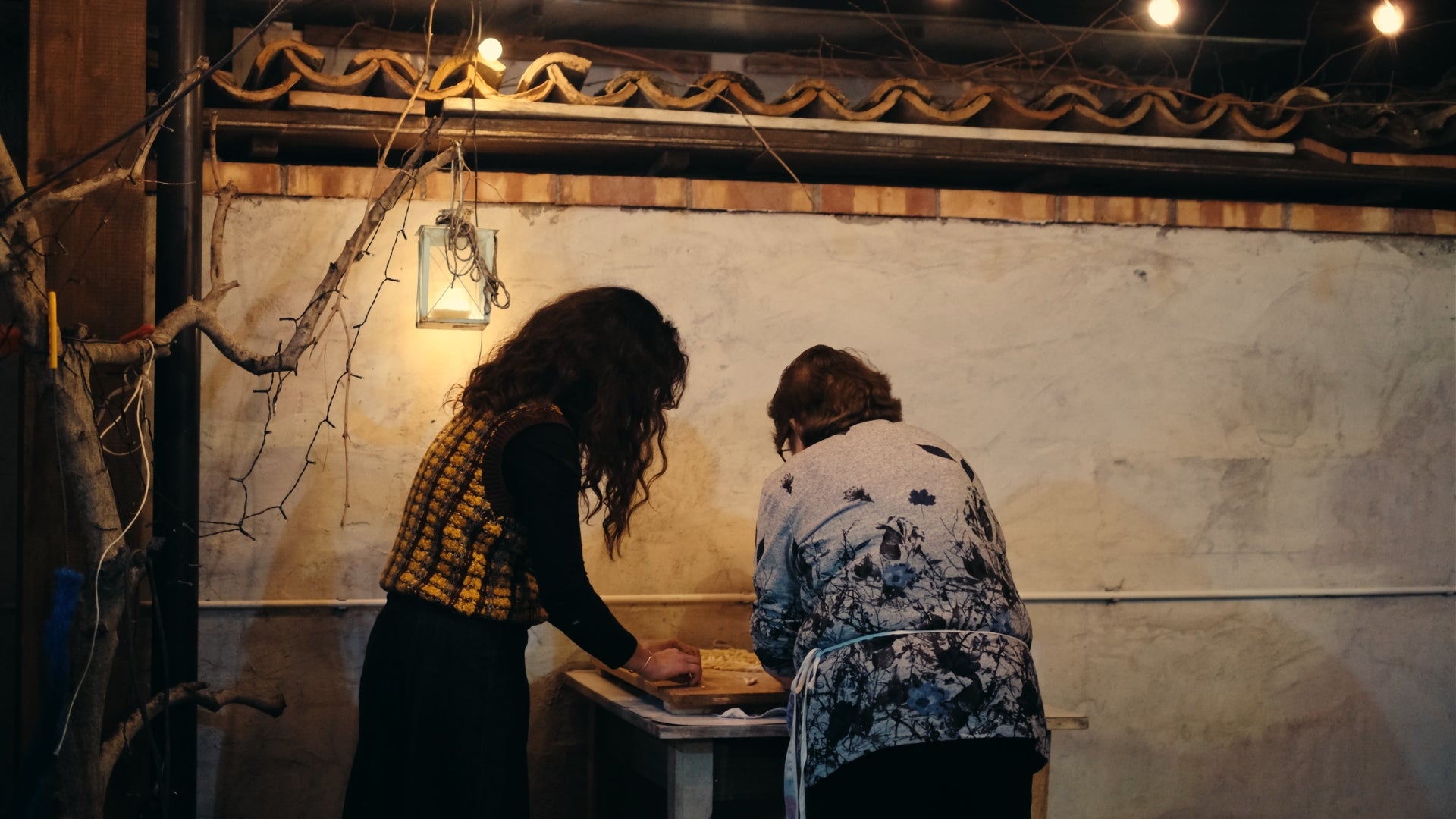 Two women stand side by side at a small table, working on something together in a dimly lit rustic room with exposed branches and string lights on the wall behind them. Their faces are not visible.