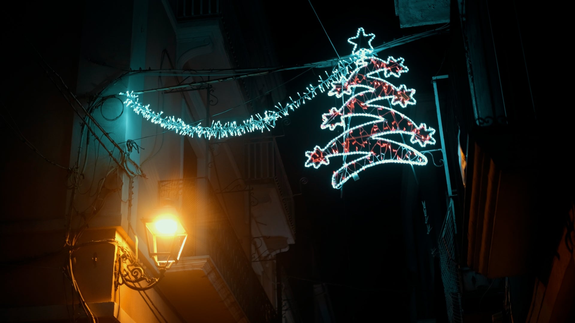 A glowing neon Christmas tree and garland hang above a narrow city street at night, illuminated by a warm street lamp below. The surroundings are dark, with balconies and building outlines visible.
