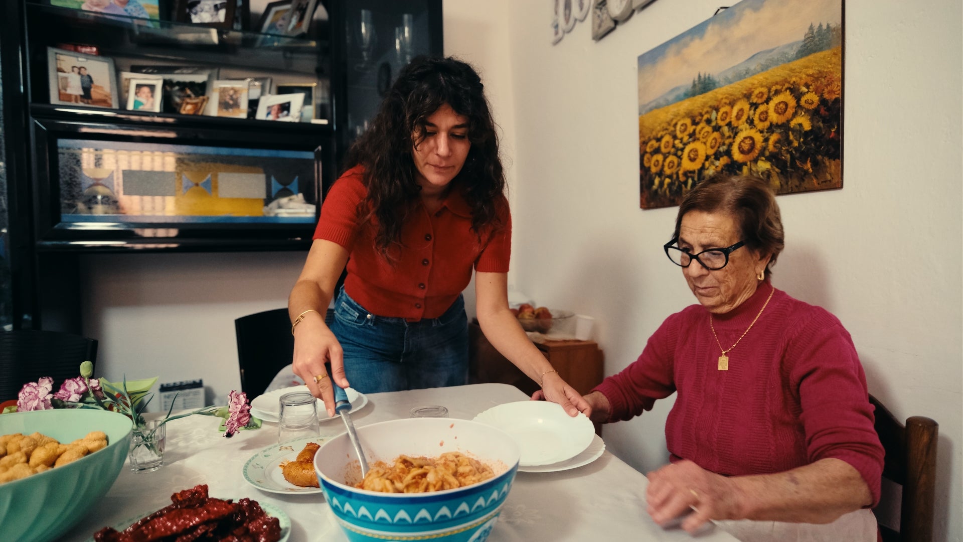 A young woman in a red shirt serves pasta from a bowl to an elderly woman wearing glasses and a pink sweater, who is sitting at a table set with food in a cozy dining room.