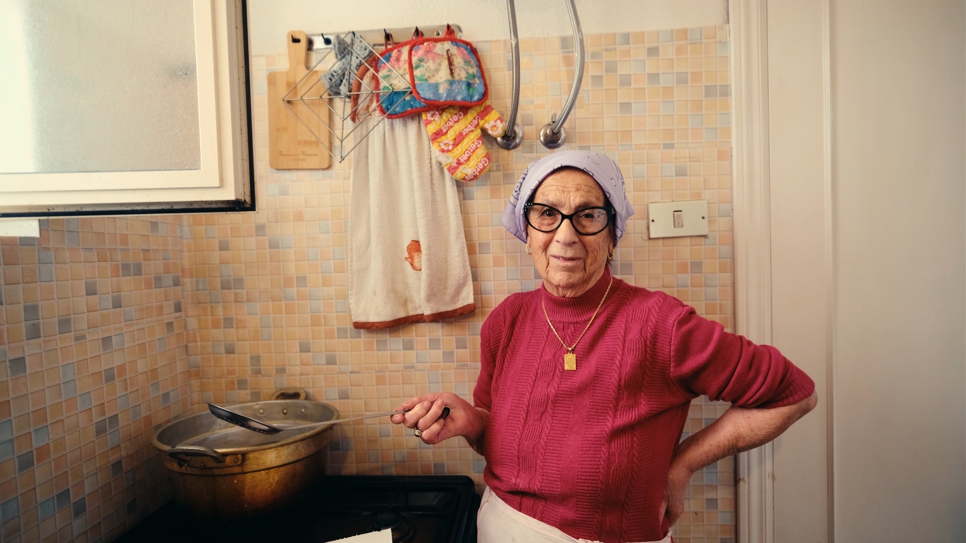 An older woman wearing glasses, a headscarf, and a red sweater stands in a kitchen, holding a cooking utensil by a stove. Colorful oven mitts and a towel hang on the tiled wall behind her.