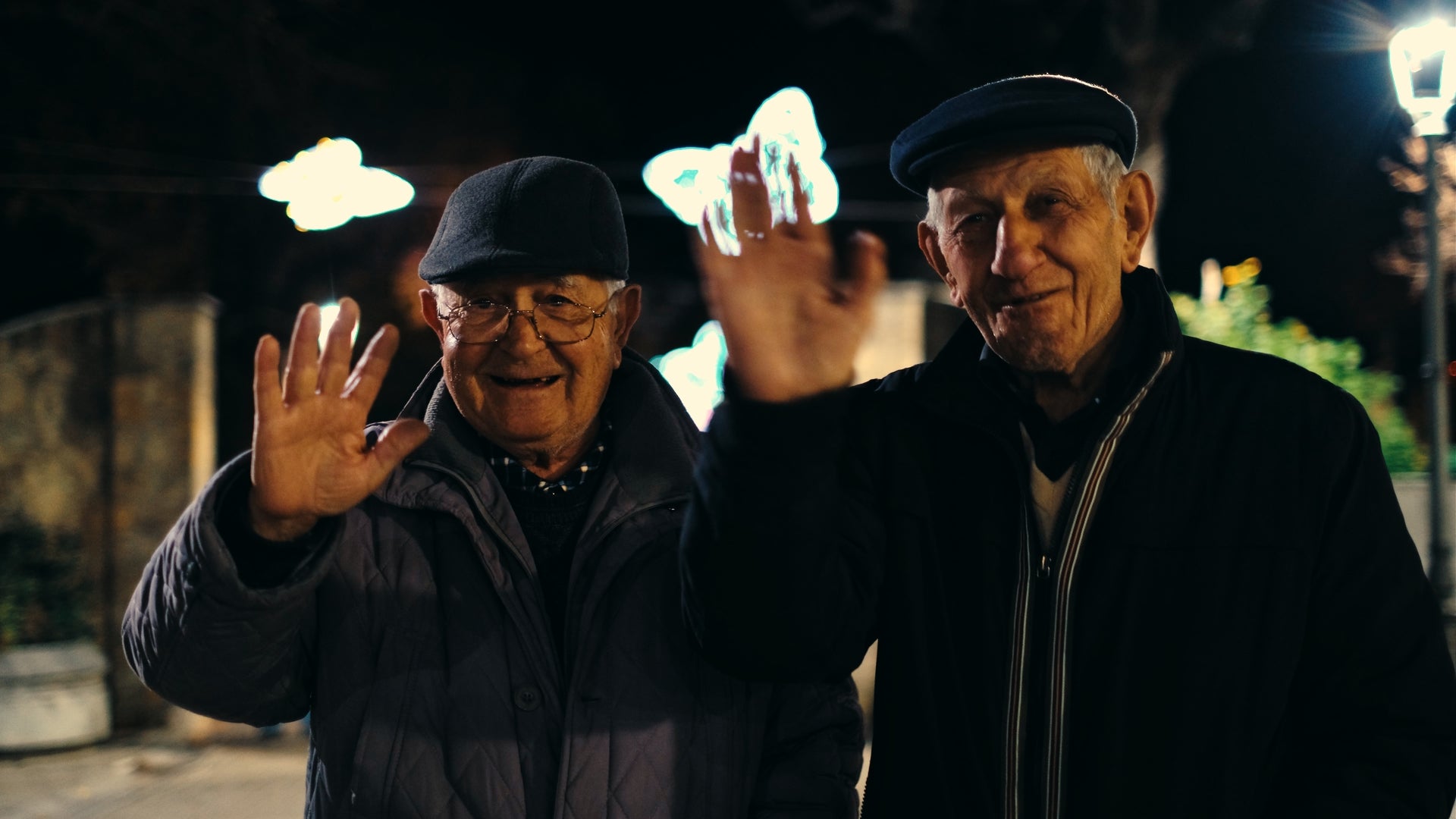 Two elderly men wearing hats and jackets smile and wave at the camera at night. Bright, decorative lights shaped like butterflies glow in the blurred background.