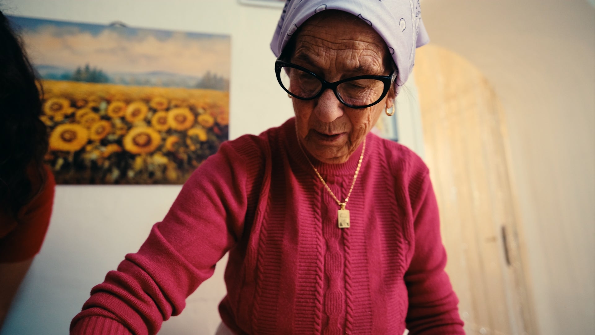 An elderly woman wearing glasses, a headscarf, and a pink sweater looks down and focuses on her task. A sunflower painting hangs on the wall behind her in a brightly lit room.