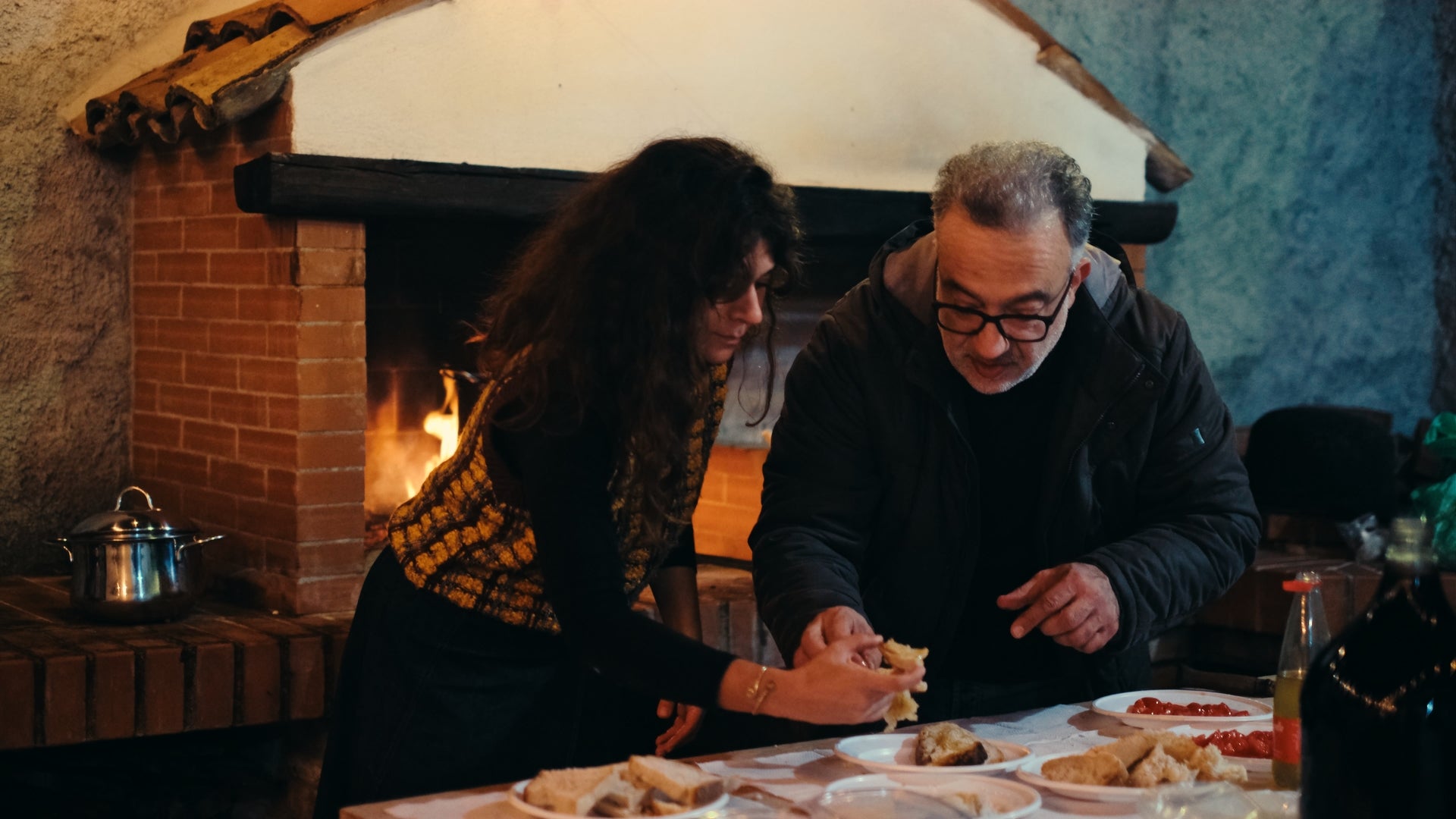 A woman and a man share food at a table set with bread and other dishes, in front of a lit fireplace in a cozy, rustic kitchen.