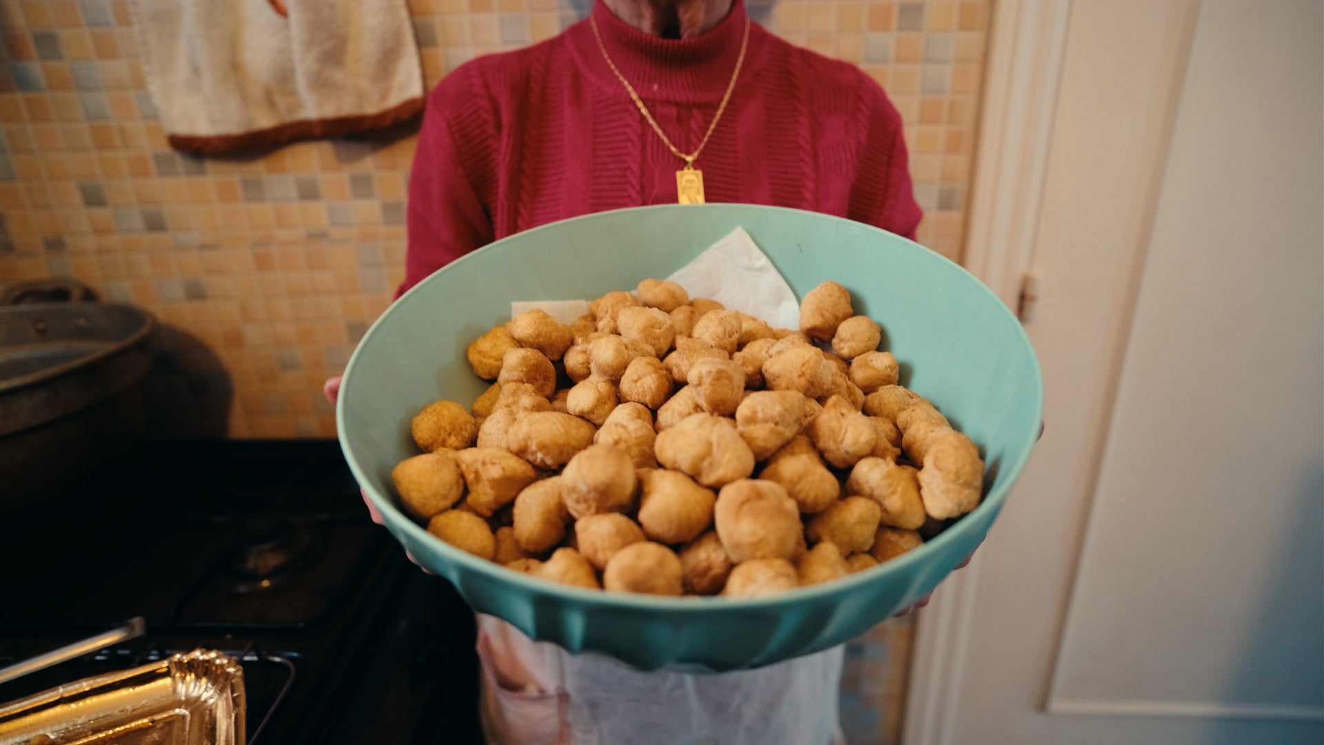 A person in a red sweater holds a large teal bowl filled with many small, round, golden-brown fried snacks in a kitchen with a tiled wall in the background.