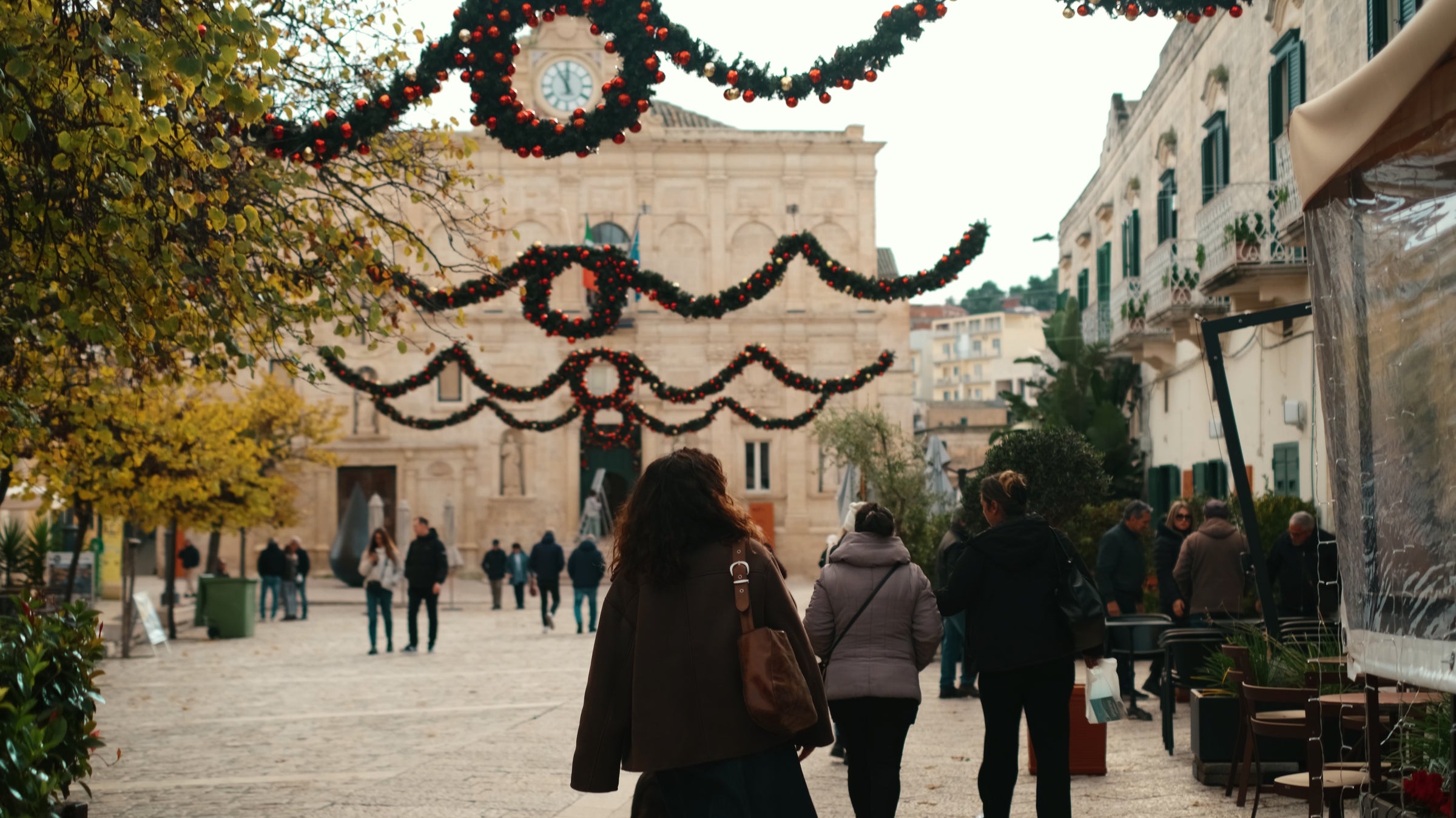 People walk through a town square decorated with Christmas garlands and red ornaments hanging overhead. A historic stone building with a clock is visible in the background. Trees line the square.