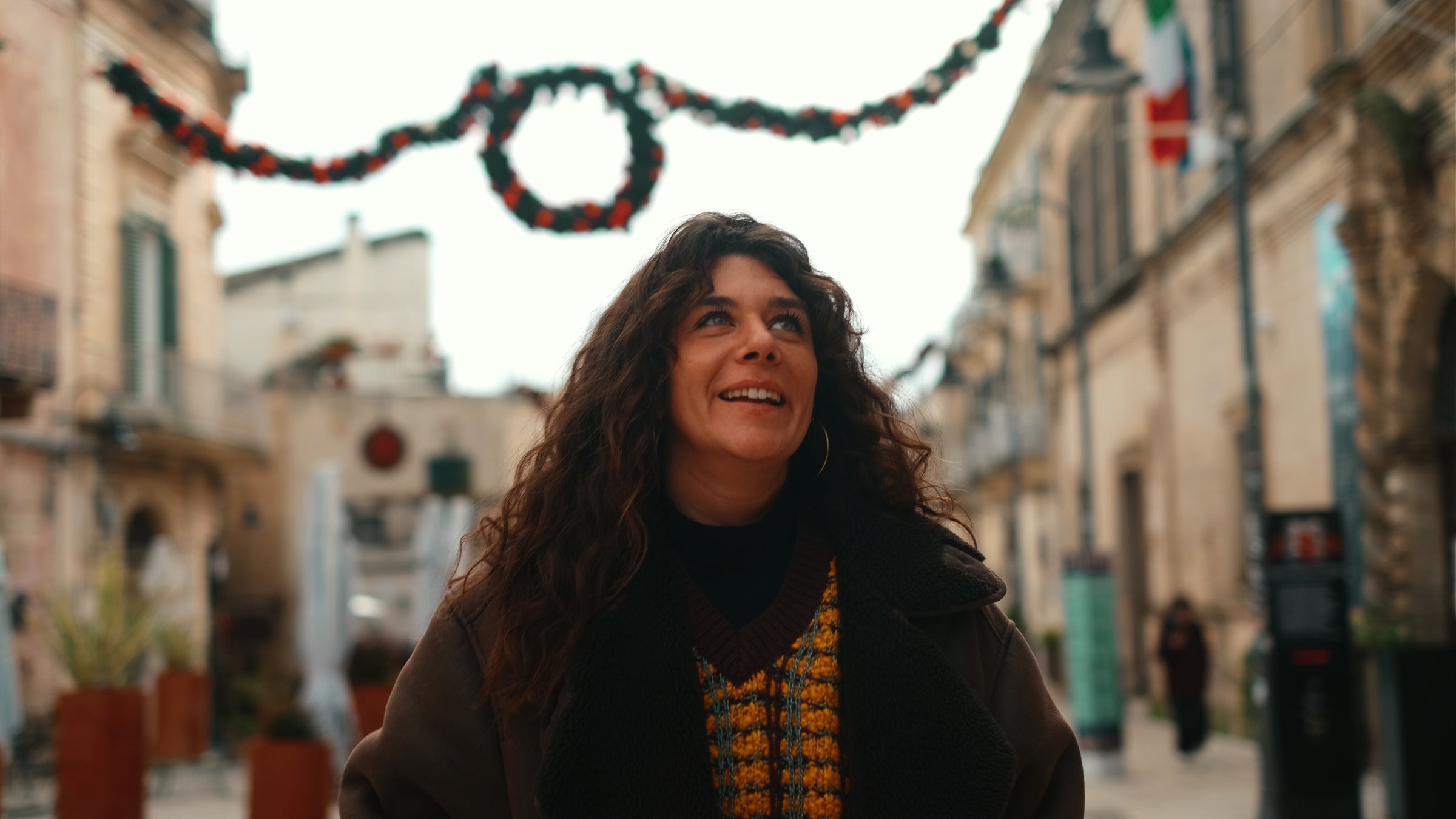 A woman with long curly hair smiles while standing on a city street decorated with festive garlands. She wears a brown coat and patterned sweater. The background is slightly blurred, highlighting the streets holiday atmosphere.