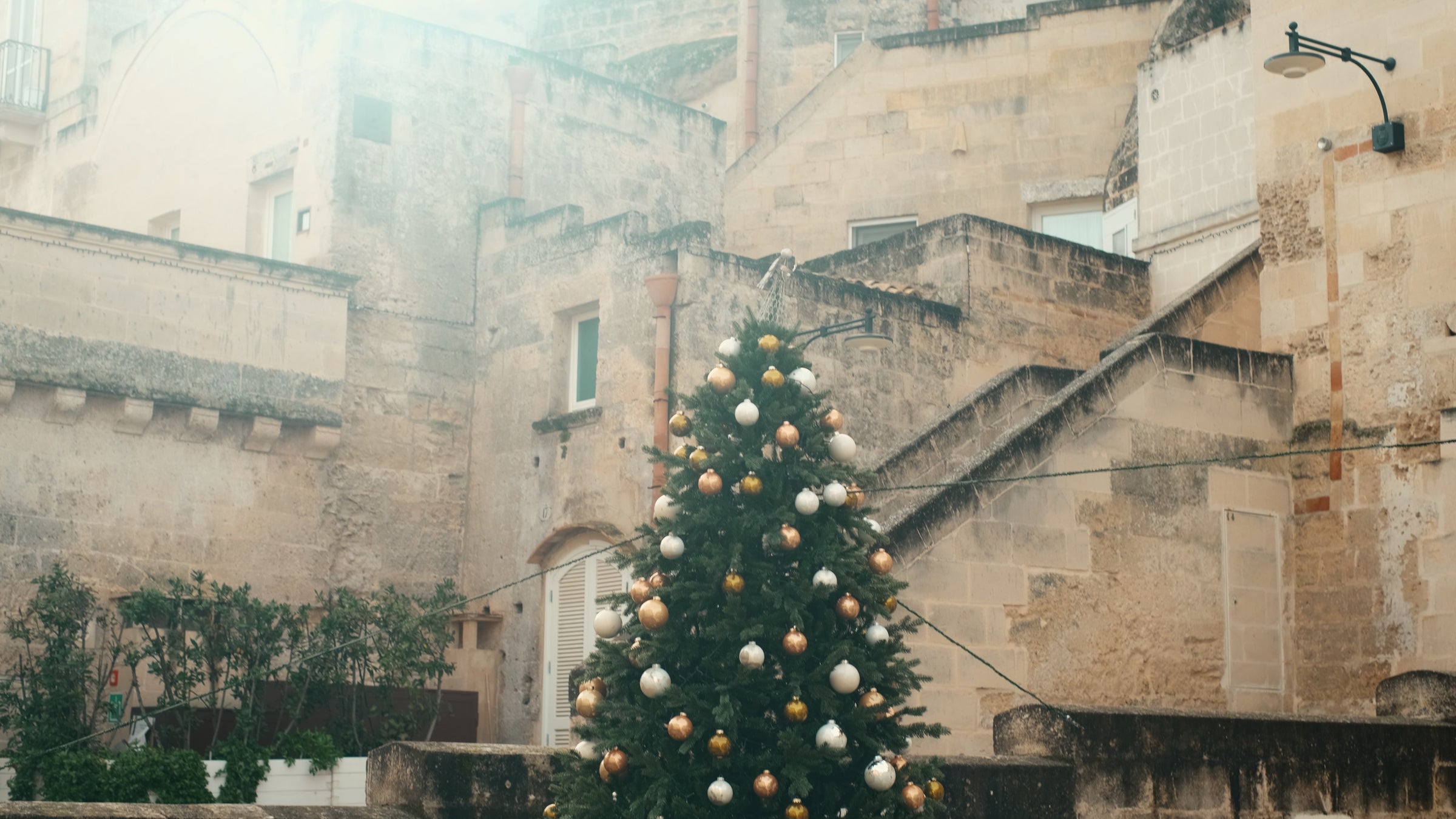 A decorated Christmas tree with white and gold ornaments stands outdoors in front of an old stone building with stairs and small windows.