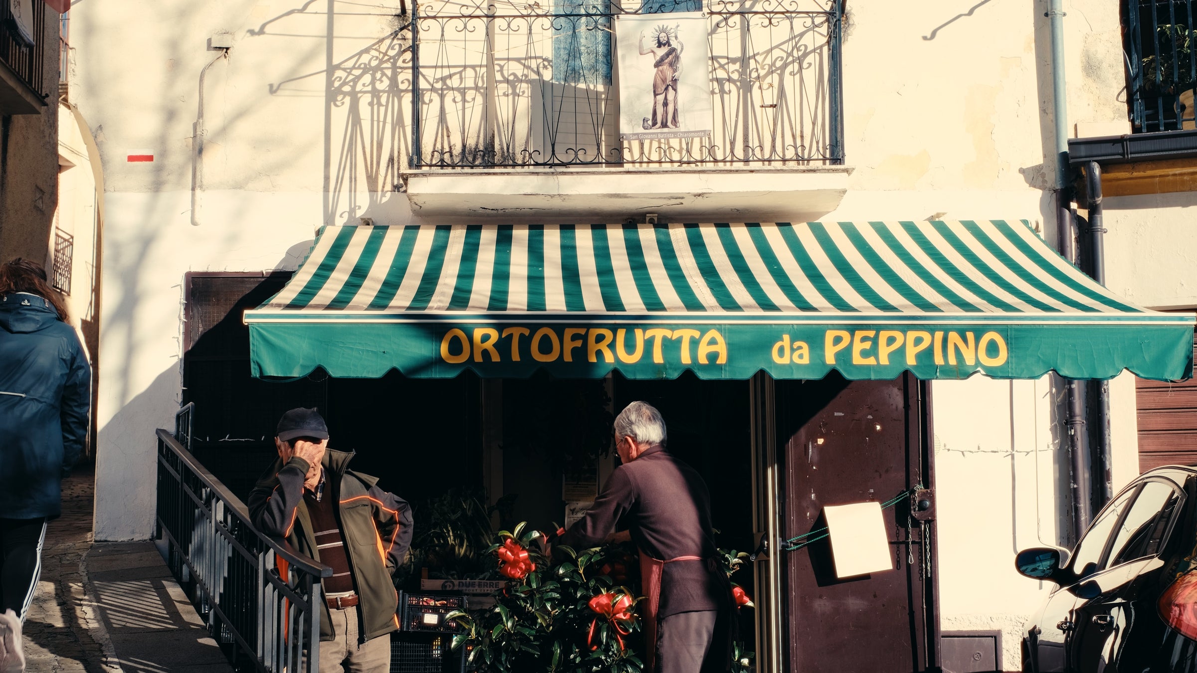 Two men stand outside a small fruit shop called Ortofrutta da Peppino with a green and white striped awning. There are plants in front, and a balcony with a painting above the shop.