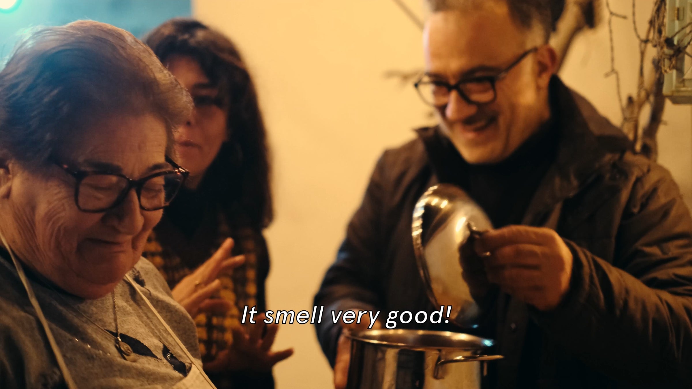 Three people smile in a warmly lit kitchen as one man lifts the lid off a large pot. The woman in front wears glasses and an apron, and the subtitle reads, It smell very good!.