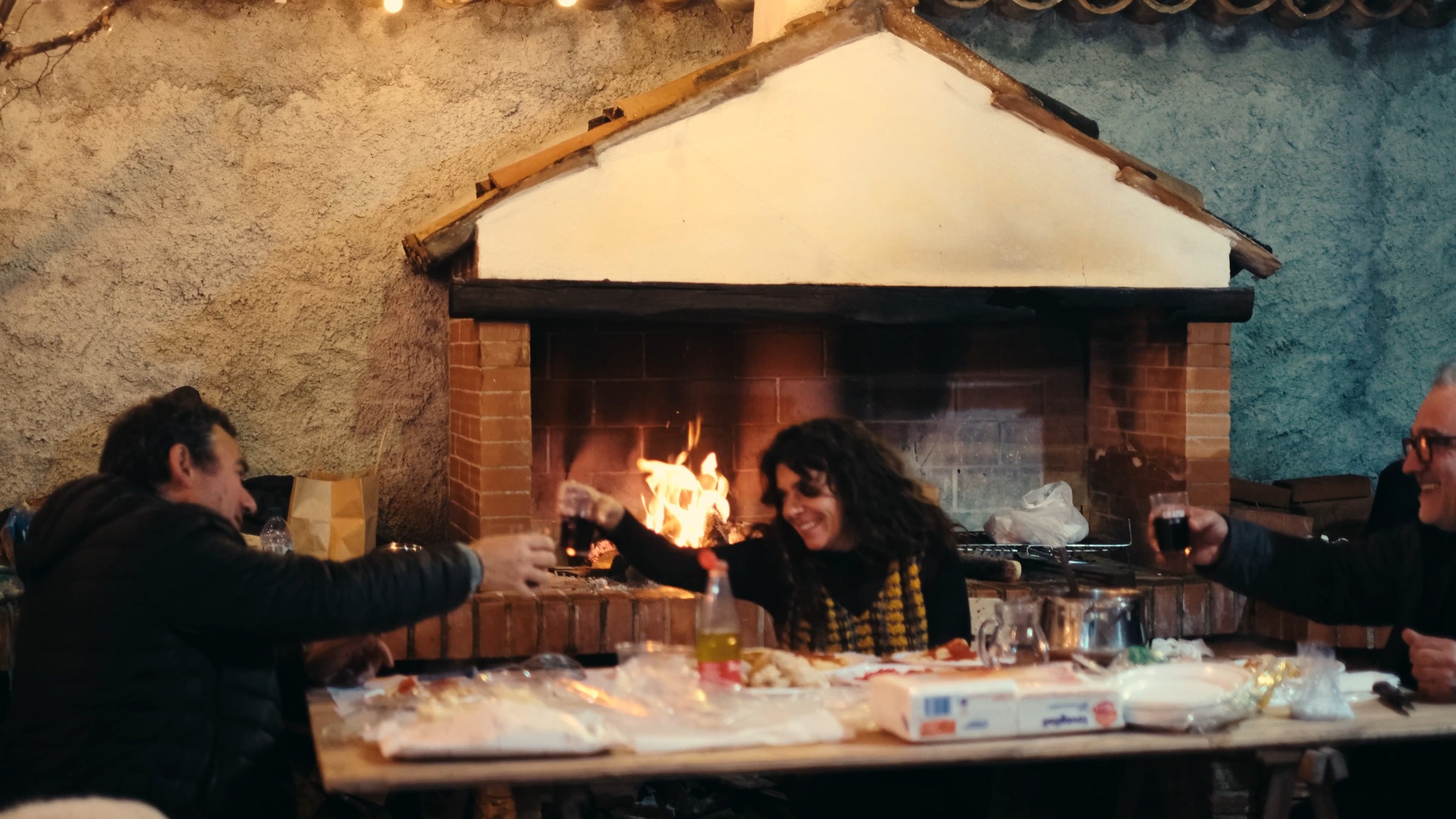 Three people sit around a table sharing a meal and clinking glasses in front of a fireplace, with food and drinks spread out and warm light creating a cozy atmosphere.