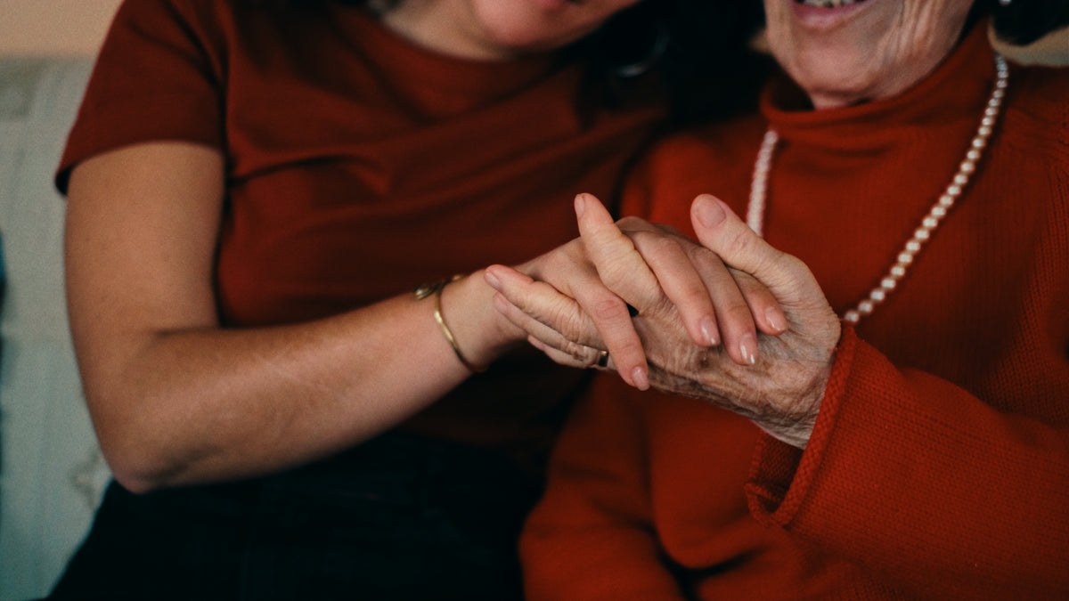 A younger person gently holds the hand of an older woman wearing a red sweater and pearl necklace, conveying warmth and connection. Both are smiling and dressed in shades of red.