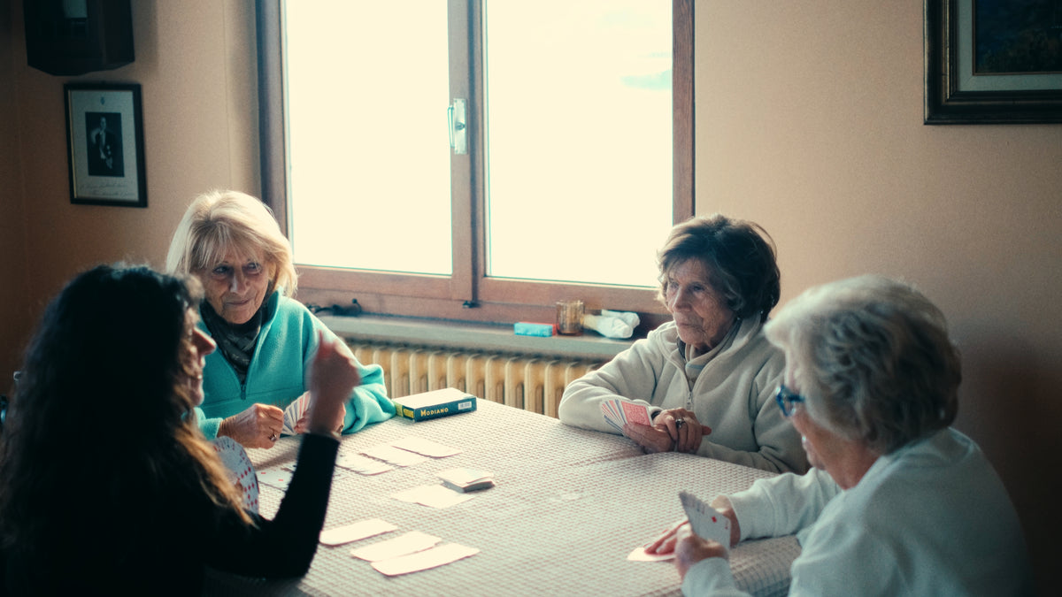 Four elderly women sit around a table playing cards in a cozy room with a large window behind them. One woman gestures while others watch and hold cards. The atmosphere appears relaxed and friendly.