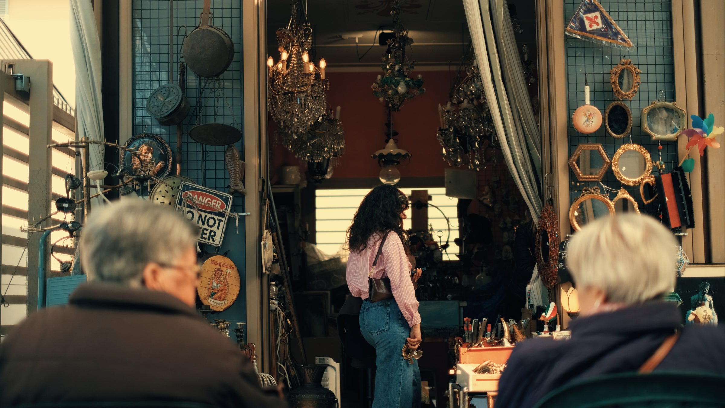 A woman with curly hair, wearing a pink shirt and jeans, stands at the entrance of a cluttered antique shop, while two people sit outside, facing the shop. Various vintage items and mirrors hang around the entrance.