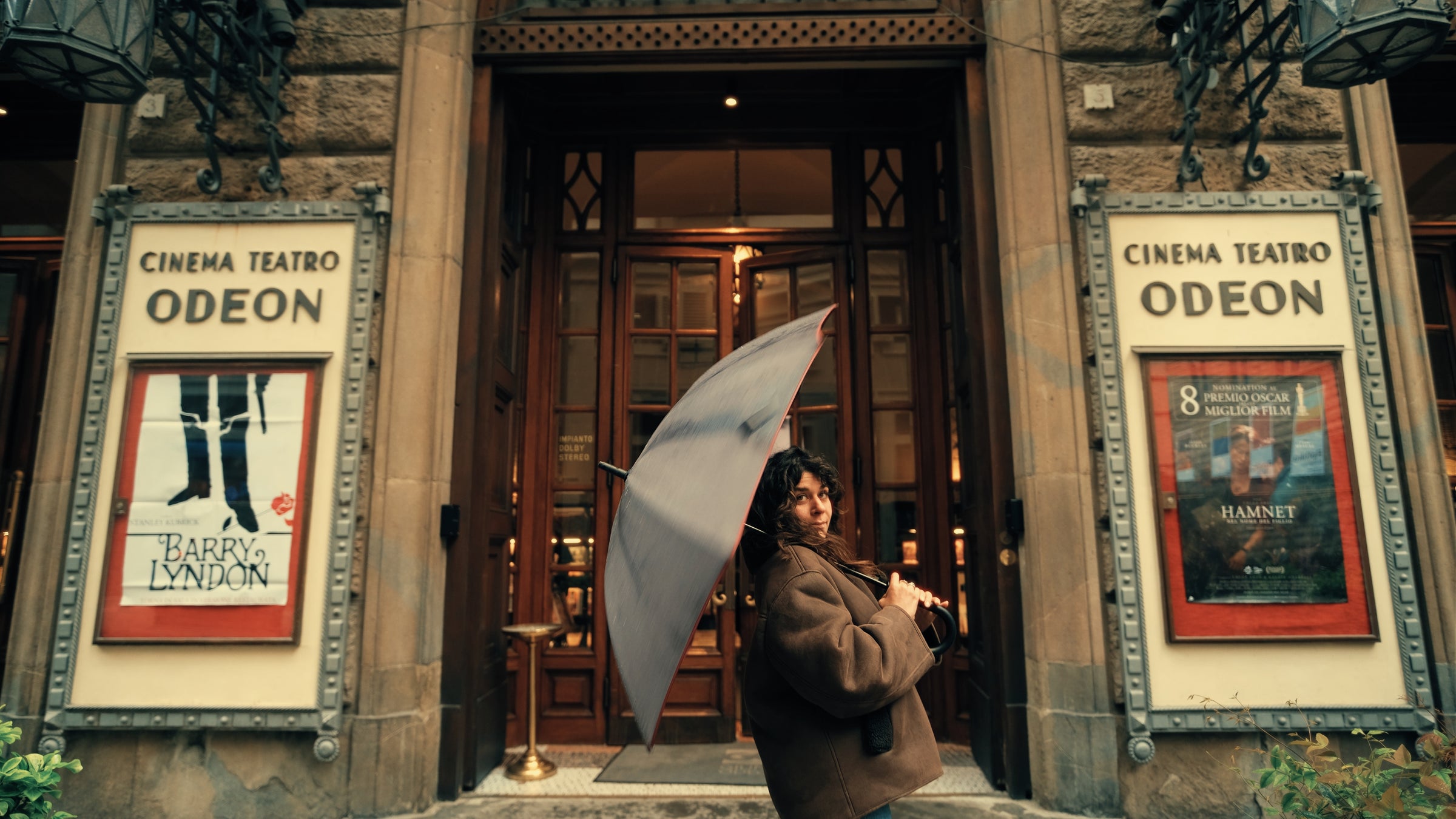 A person holding an umbrella stands in front of the ornate entrance of Cinema Teatro Odeon, with vintage movie posters displayed on either side of the doorway.