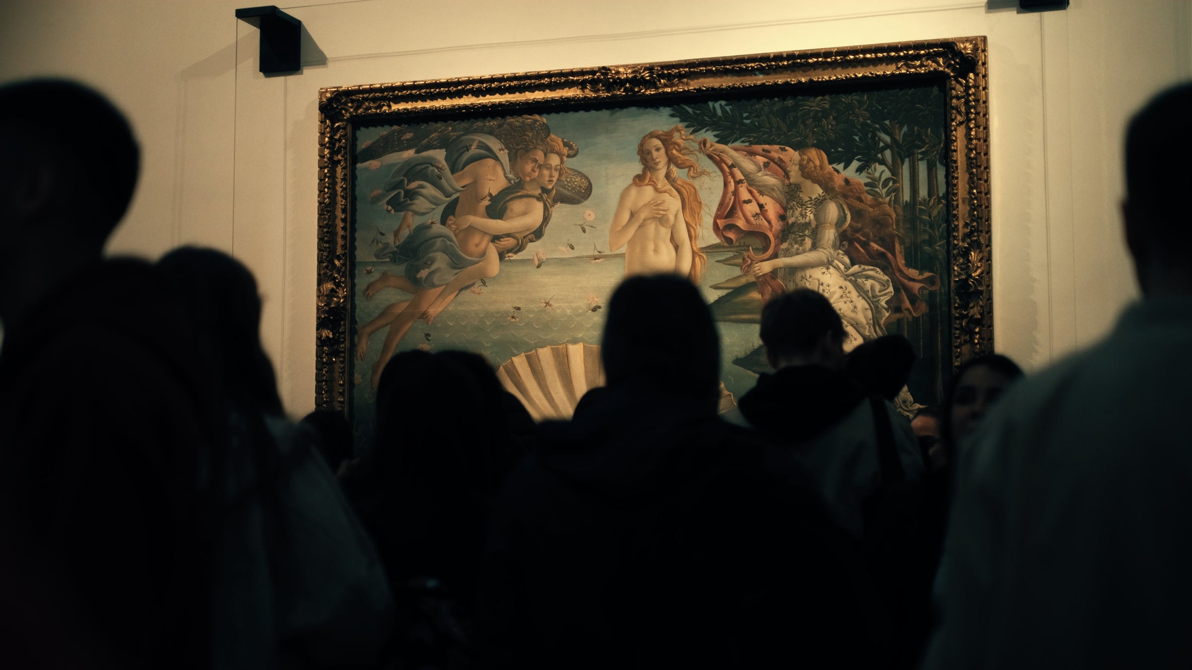 A group of people stands in front of Sandro Botticelli’s famous painting The Birth of Venus, which is framed and displayed on a museum wall. The crowd’s silhouettes are dark against the lit artwork.