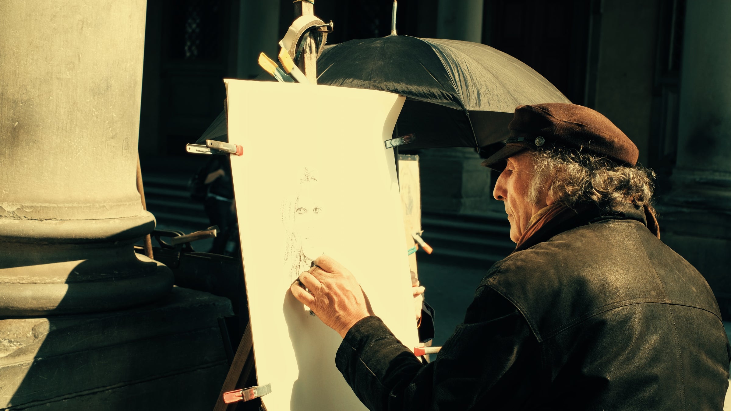 An artist wearing a brown cap sketches a portrait on a large white sheet, using a pencil, outdoors. Sunlight highlights his face and hand, and an umbrella is positioned behind the drawing for shade.