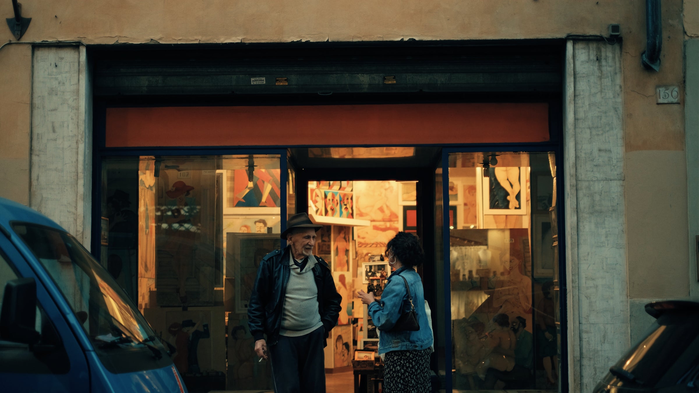 An elderly man and a woman stand talking outside an art gallery with warm lighting and colorful paintings visible through the large front windows. A blue car is parked in front of the gallery.