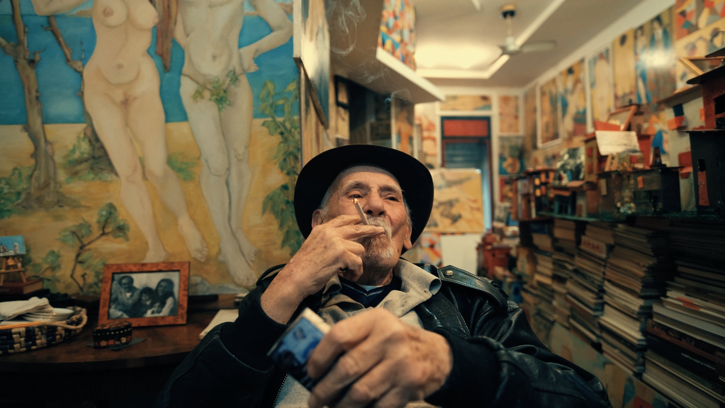 An elderly man wearing a hat and leather jacket sits in a colorful, art-filled room, holding a cigarette. Behind him are numerous paintings, including nude figures, as well as stacks of books and framed photos.