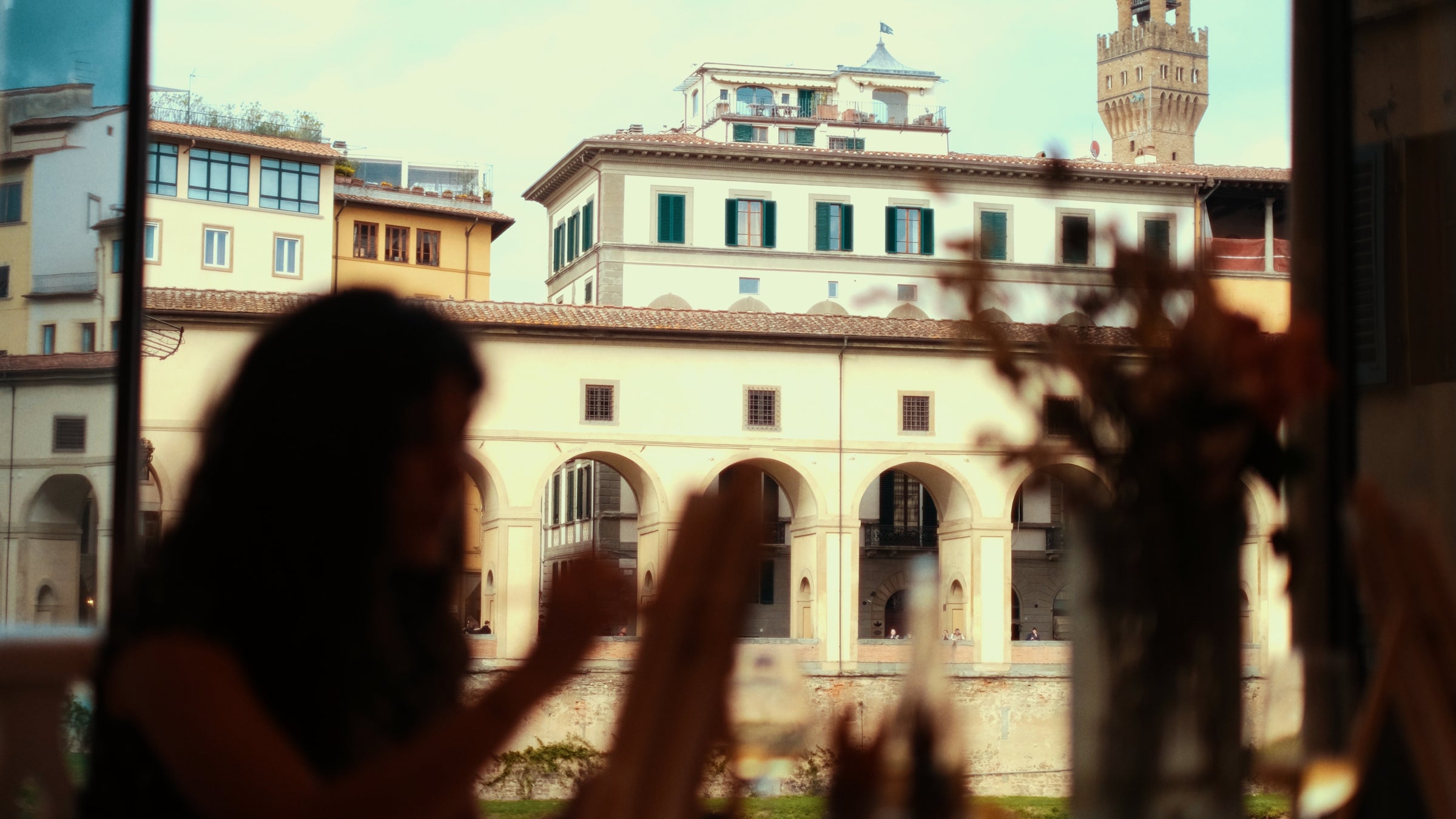 A silhouette of a person dining indoors, with a view of historic buildings and arched architecture outside through a large window, likely in an Italian city.
