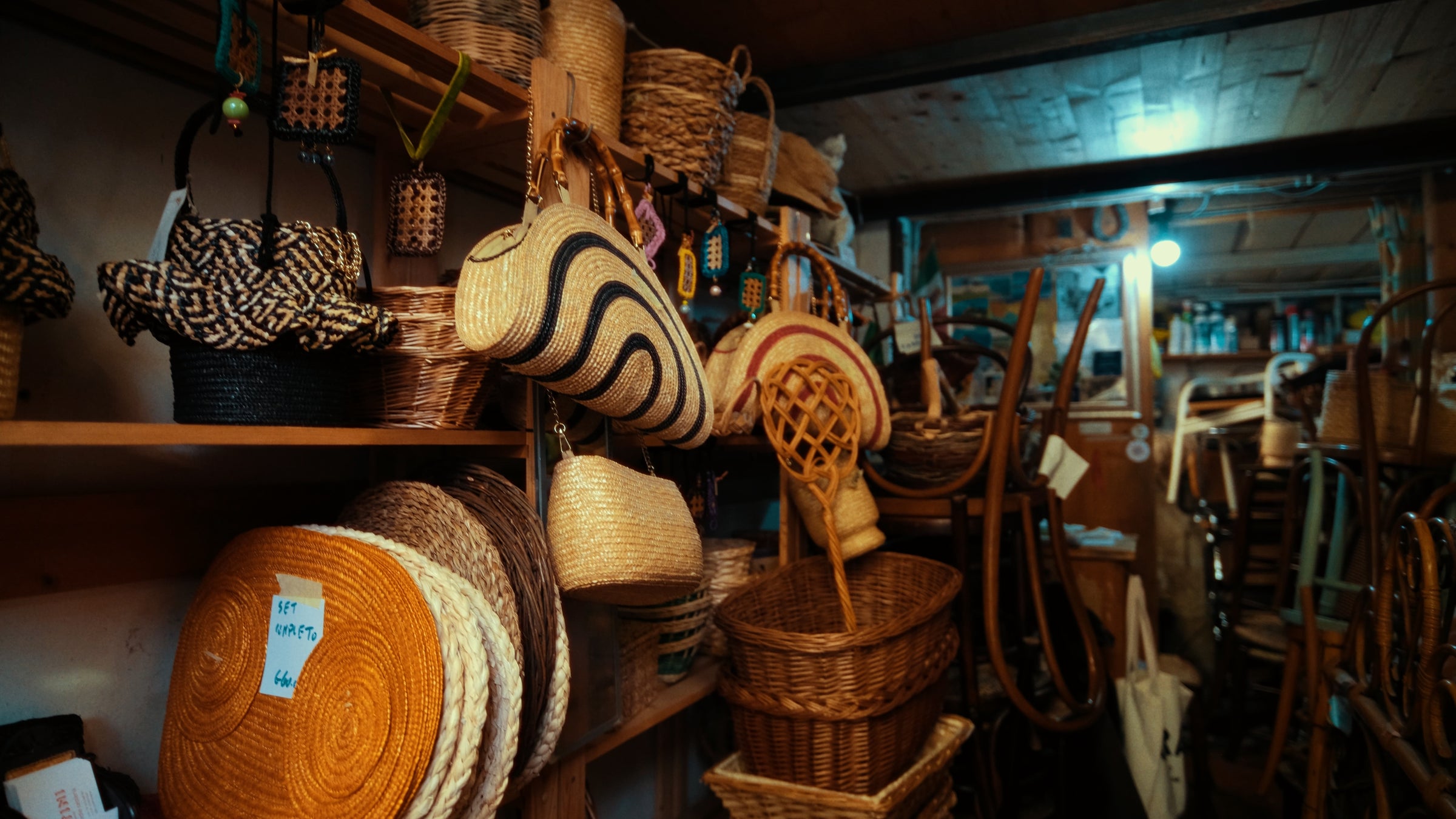 A cluttered shop interior with shelves full of woven baskets and straw bags in various shapes, sizes, and patterns, alongside stacked wicker furniture and other handmade crafts under warm lighting.