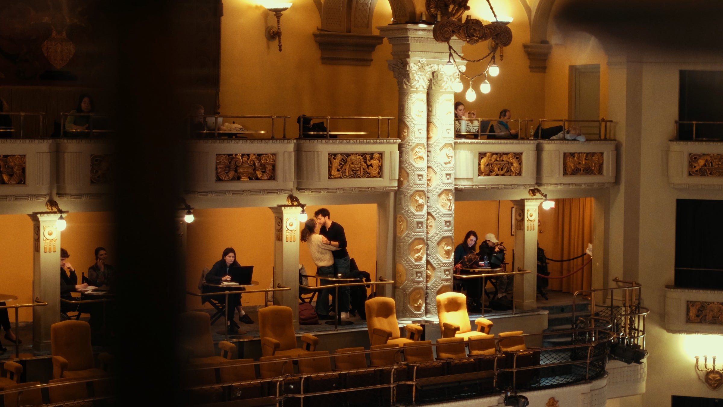 People sit at tables and work or socialize on balconies inside an ornate, warmly lit theater or concert hall. A central decorated pillar and plush seating are visible, creating a cozy, elegant atmosphere.