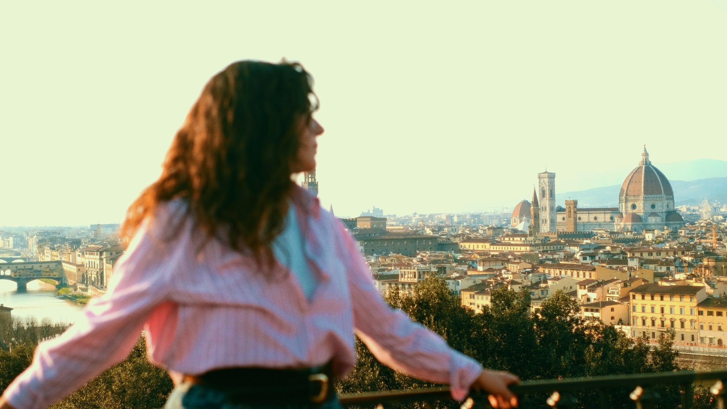 A person with wavy hair and a striped shirt stands in the foreground, blurred, overlooking the cityscape of Florence, Italy, with the Florence Cathedral and historic buildings in the background.