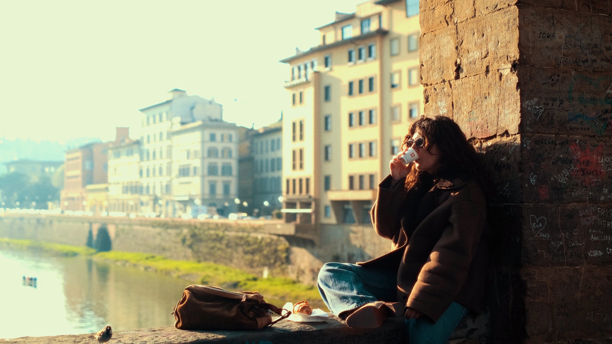 A person with curly hair, wearing sunglasses and a coat, sits by a stone wall overlooking a river, drinking from a cup. A brown bag and some food rest beside them; tall buildings line the opposite riverbank.